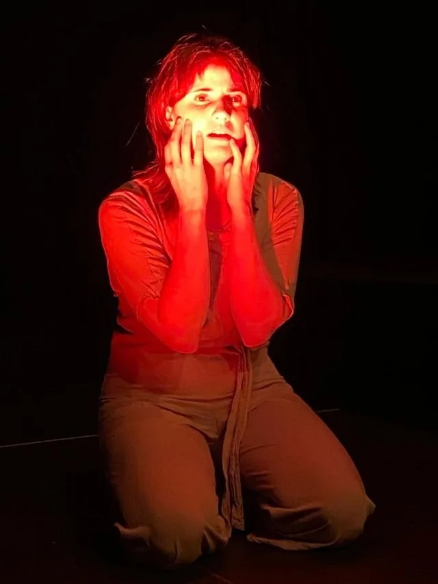 A woman with short, curly hair sitting on her knees, illuminated by red light, holding her face with both hands, appearing distressed or surprised.
