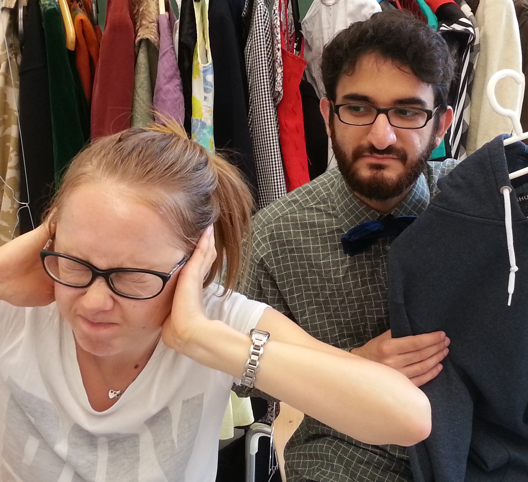 A woman in glasses holding her head with a distressed expression, next to a man with glasses, a beard, and a bow tie, looking at her in concern, in a clothing store with hangers of clothes in the background.
