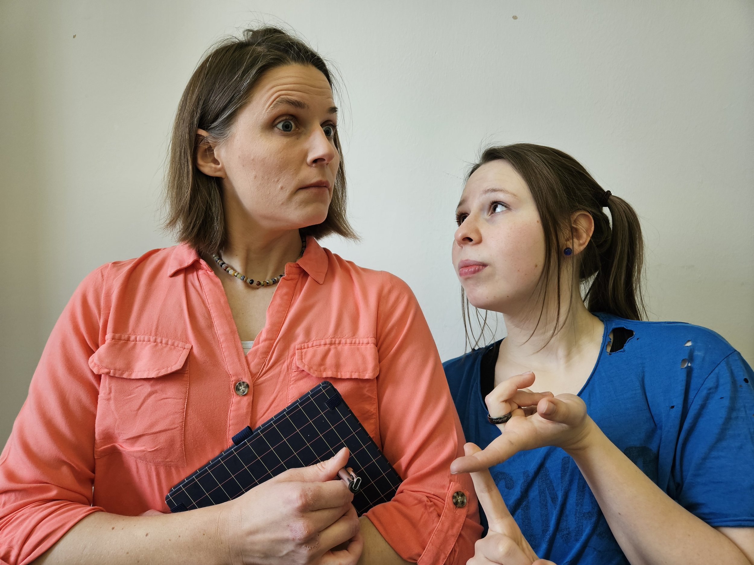 Two young women having a serious conversation. One is holding a clipboard and wearing an orange shirt, the other wearing a blue shirt with torn sleeves, making a point with her hand.