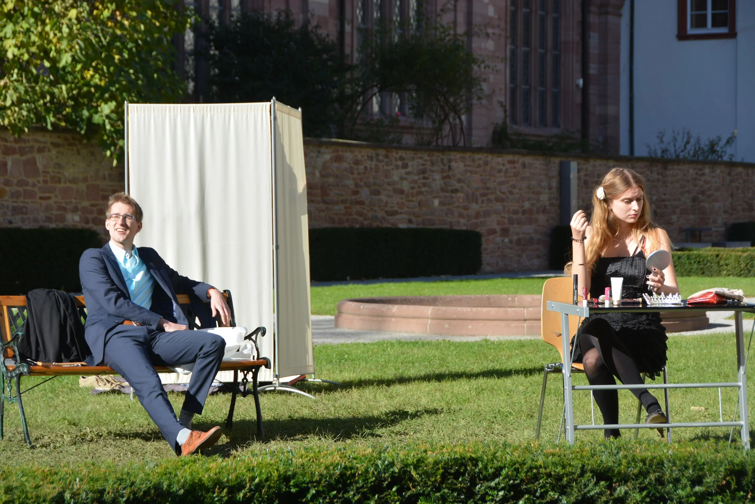 A man in a suit sitting on a park bench and a woman with makeup at a table applying makeup outdoors on a sunny day.