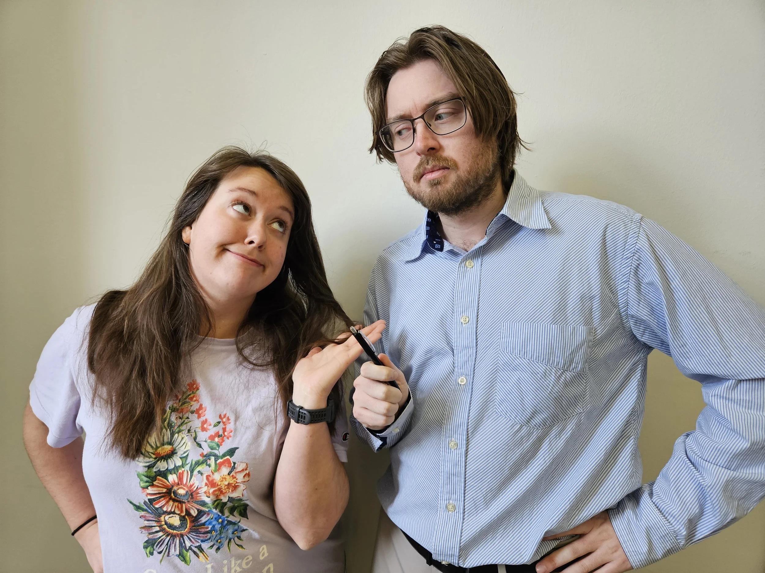 A woman with long brown hair smiling playfully with her eyebrows raised and looking at a man. The man has shoulder-length brown hair, glasses, and a beard, and is looking at the woman with a slightly skeptical or unimpressed expression. The woman is wearing a white t-shirt with a colorful floral design, and the man is wearing a light blue, striped, button-down shirt. Both are standing against a plain, light-colored wall.