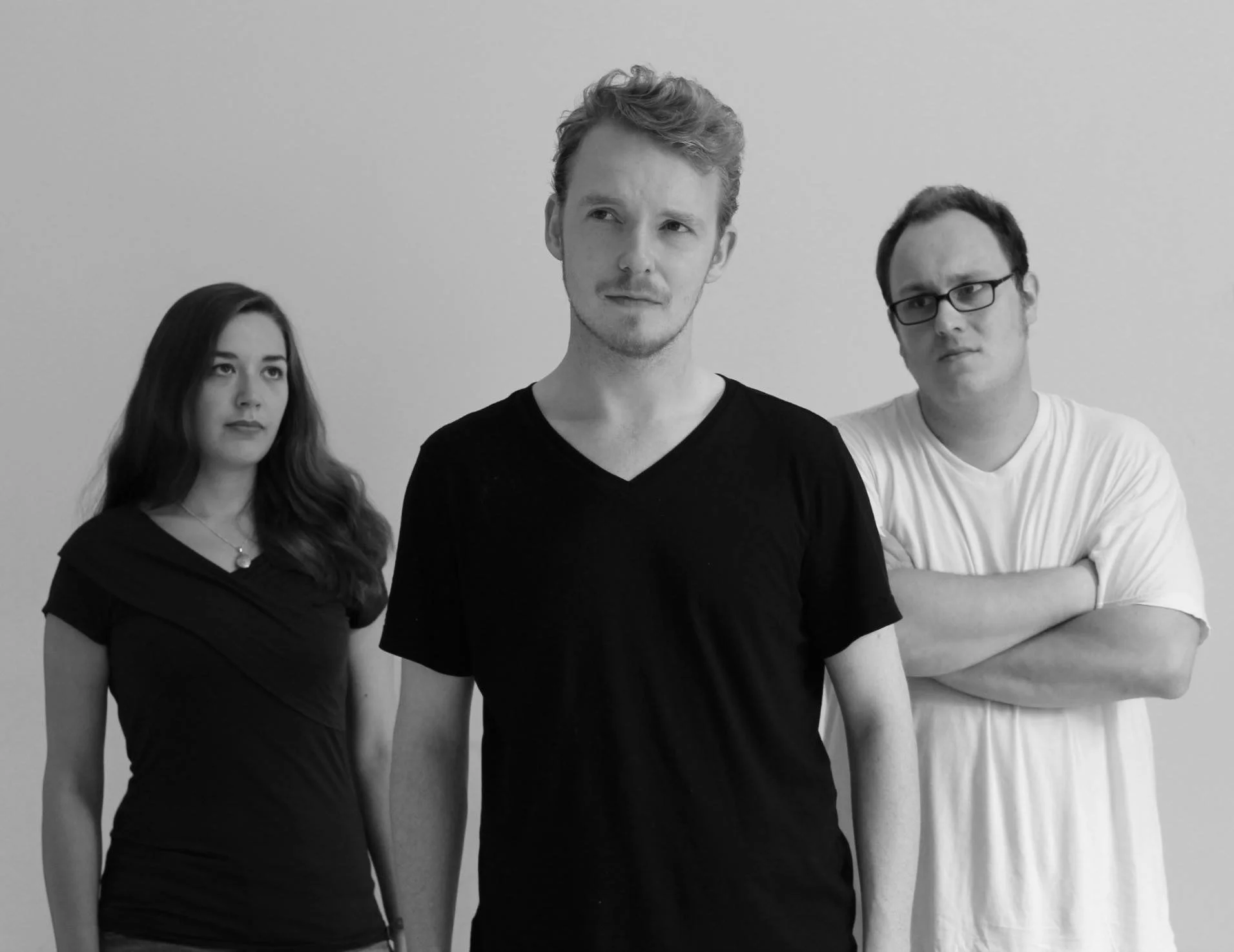 Three young adults standing against a plain gray wall. A woman with long wavy hair and a necklace on the left, a man with curly hair in the middle, and another man with glasses and short hair on the right. All appear serious and focused.