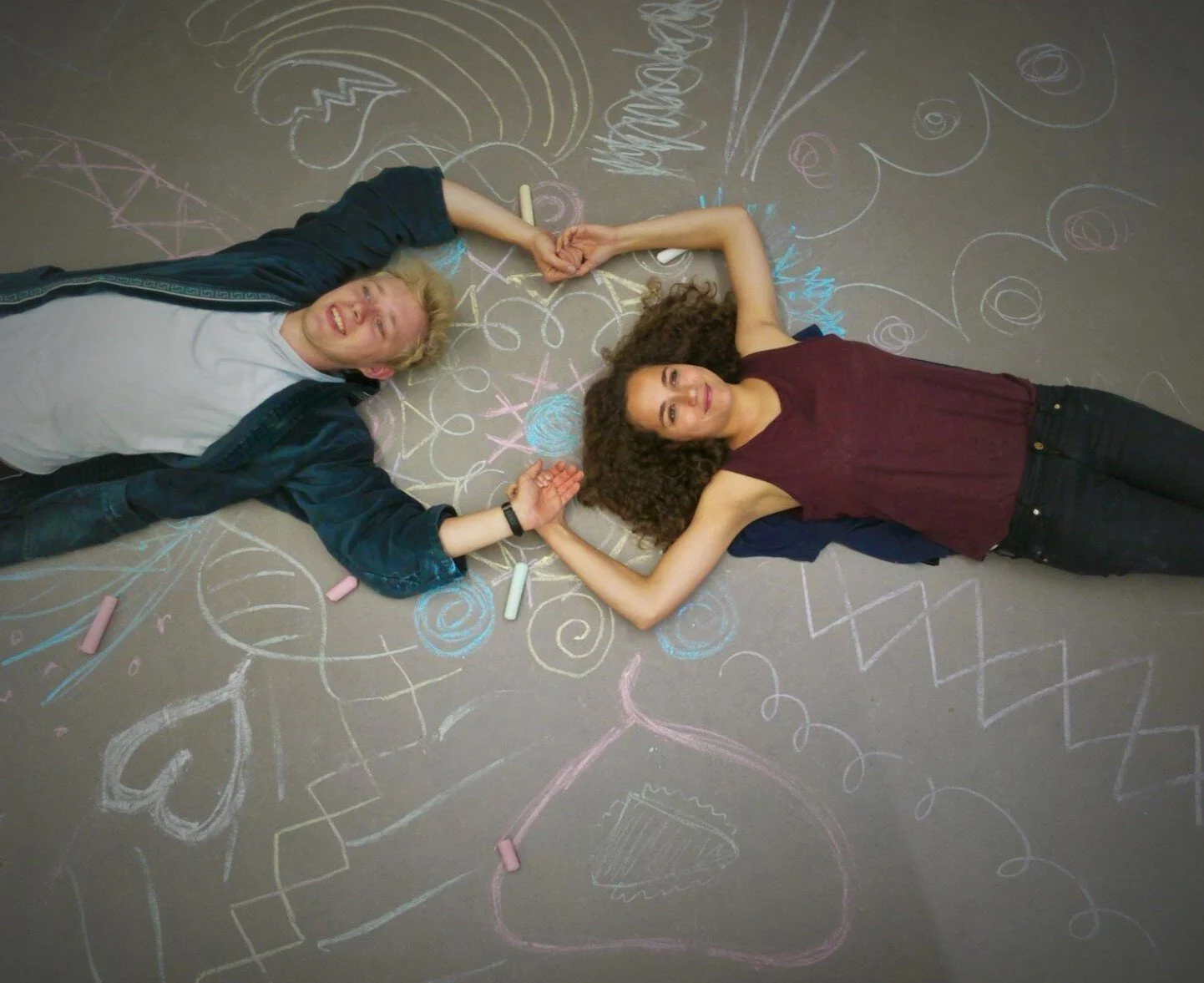 Two young women lying on a chalkboard floor, surrounded by colorful chalk drawings, holding hands and smiling.