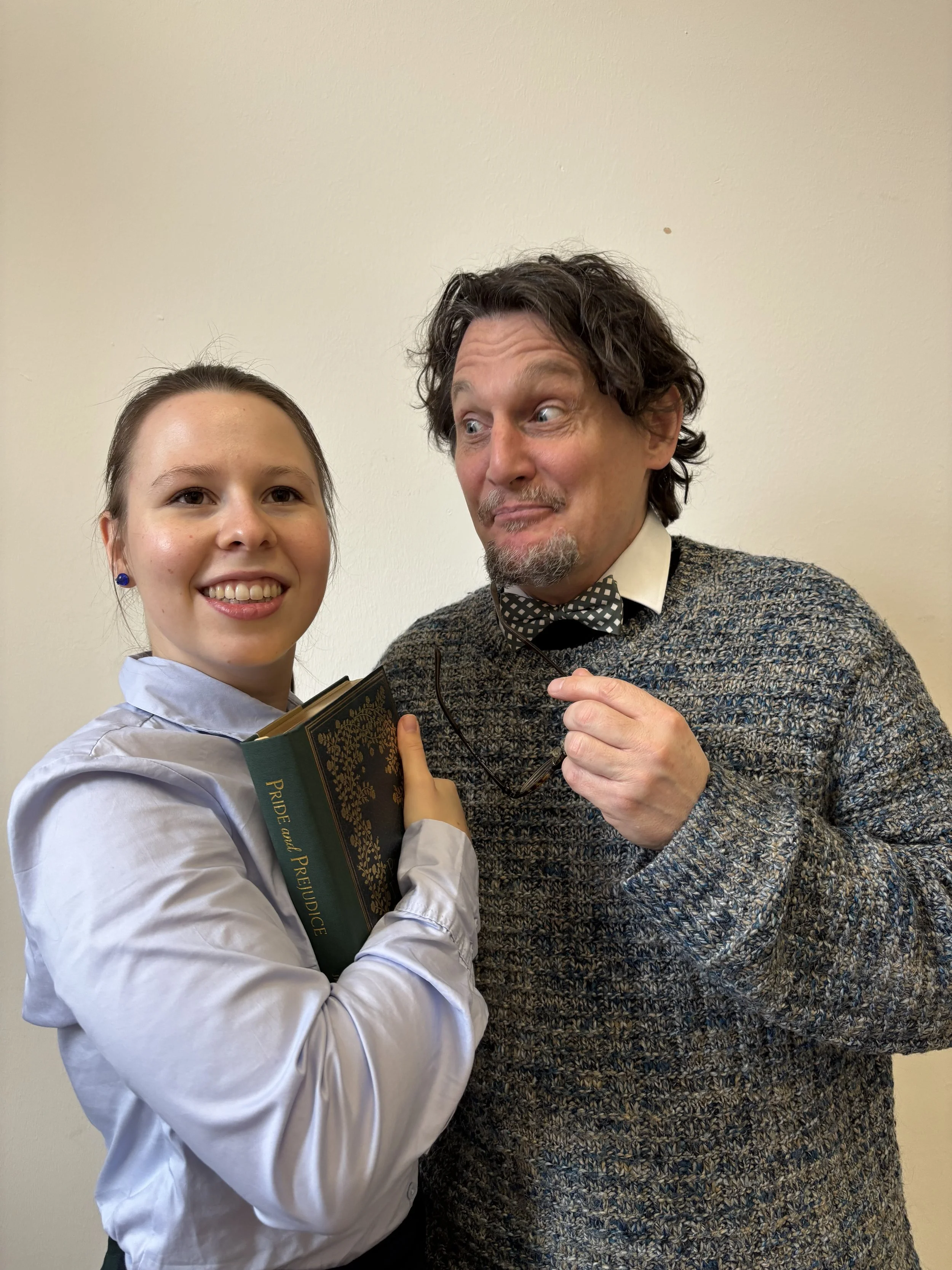 A young woman holding a book titled 'PRIDE and PREJUDICE', standing next to a man with curly hair holding glasses, both smiling and looking at each other against a plain wall.