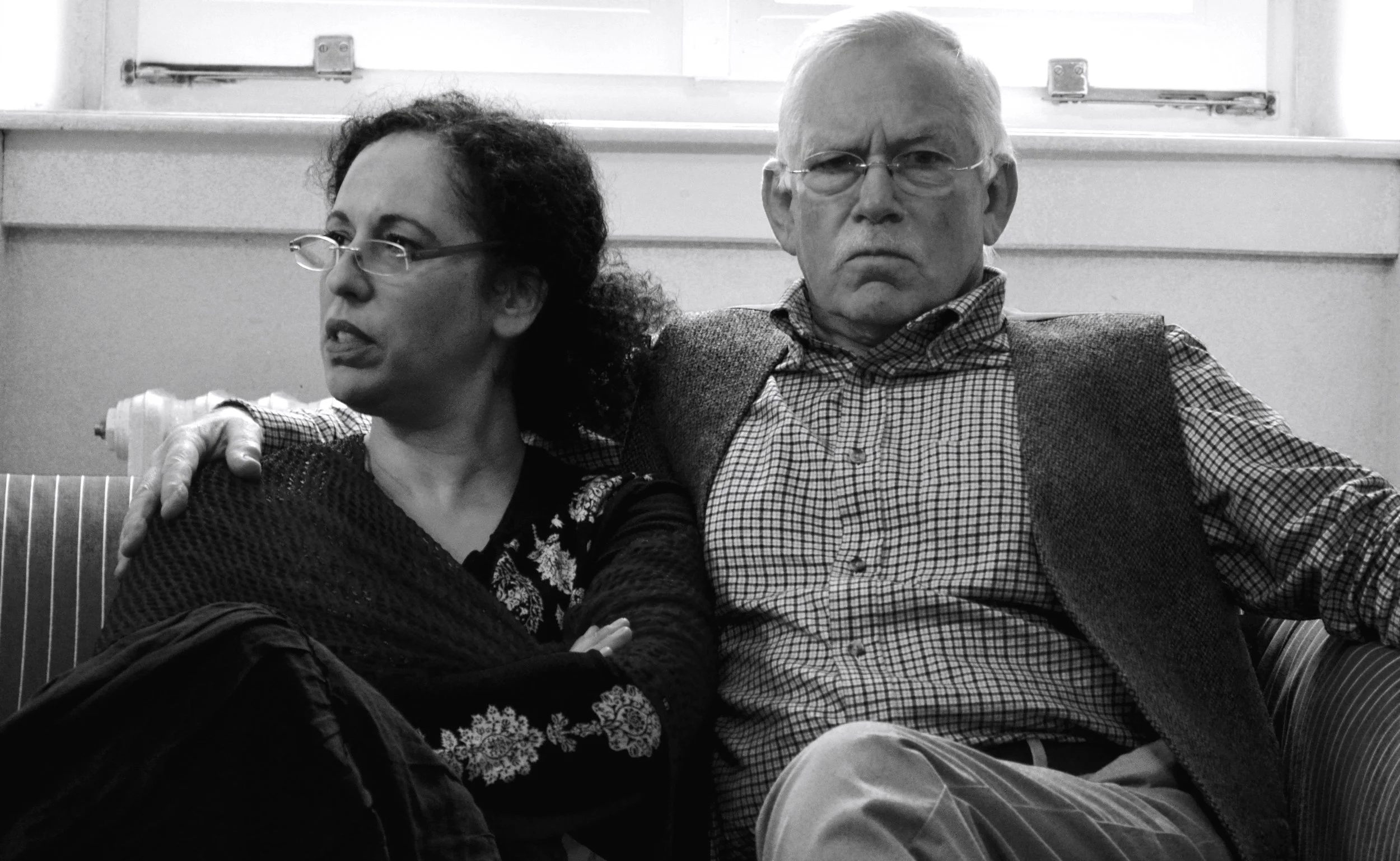 A black and white photo of a woman and an older man sitting side by side on a striped sofa. The woman has curly hair, wears glasses, and a dark sweater. The man has glasses, a checked shirt, and a vest. They both look serious, with the woman looking to the side and the man directly at the camera.