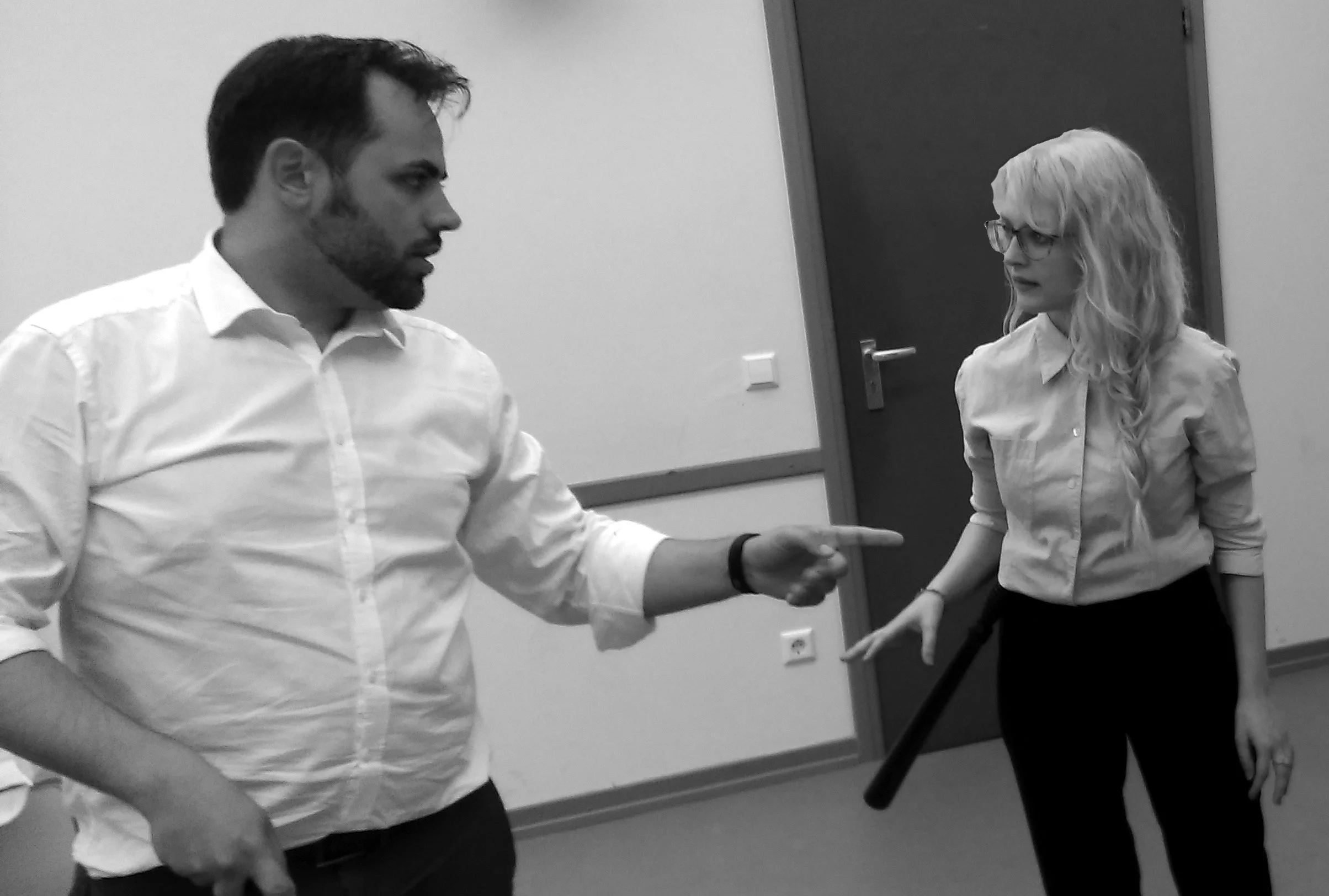 A man pointing at a woman while talking inside an office, both dressed in formal attire.