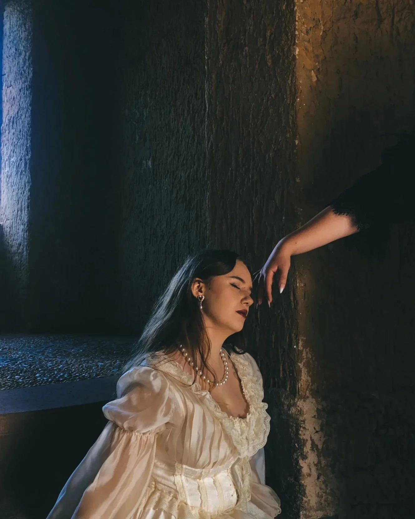 A woman in vintage clothing with pearl necklace and earrings, posing with her eyes closed in a dark, textured room, as someone gently touches her forehead.
