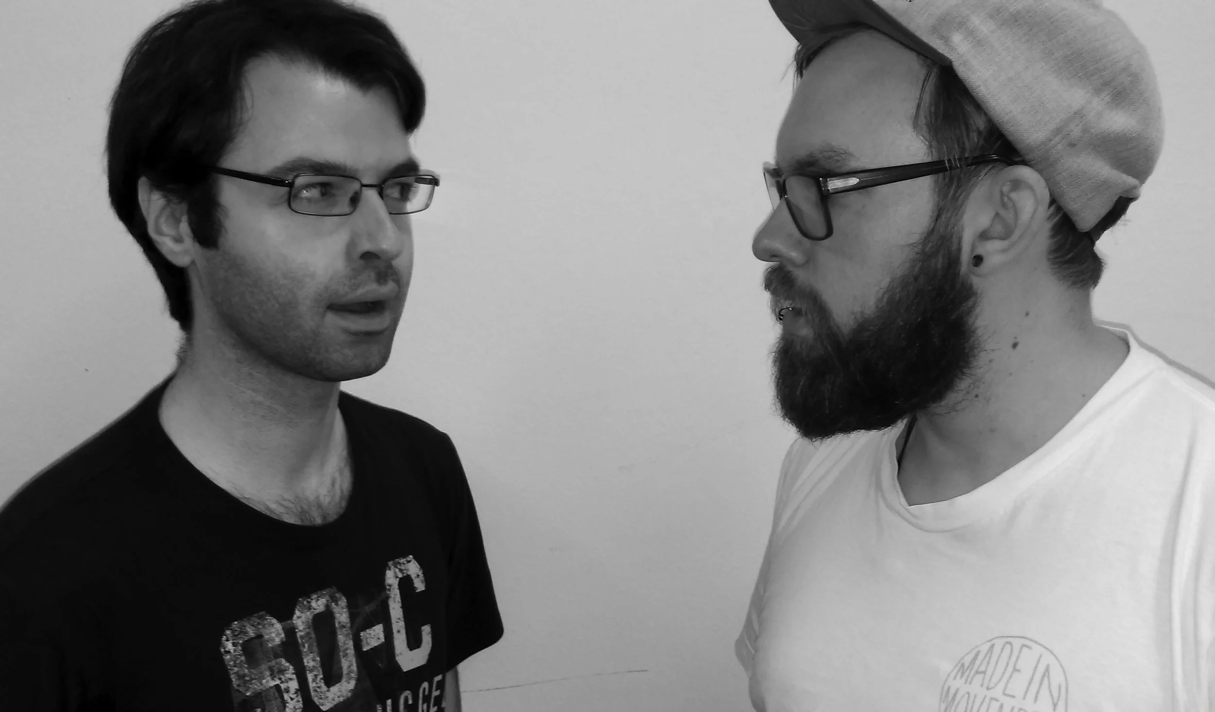 Two men with glasses and beards are facing each other and talking against a plain wall.