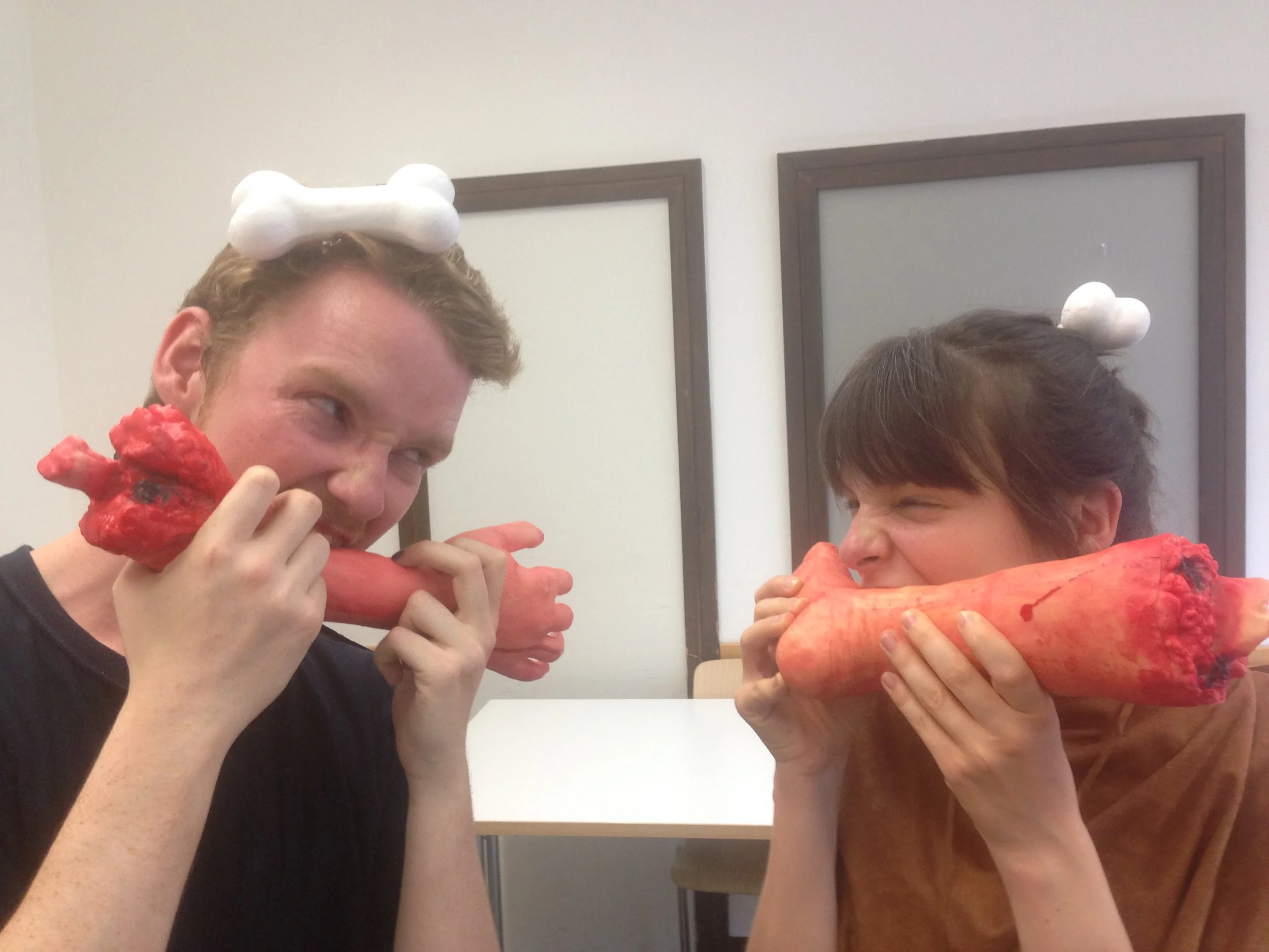 Two teenagers with headbands topped with paper bones, pretending to bite large, fake, red and orange plush bones, sitting in a room with white walls and benches.