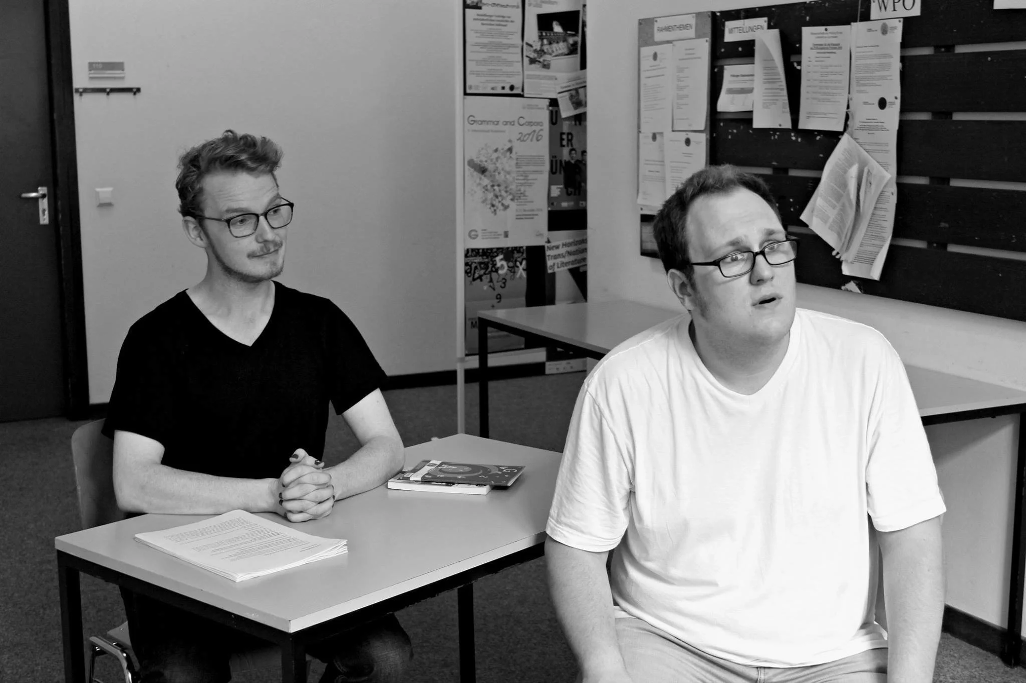 Two men sitting at a table in a classroom or office setting, one with glasses and a black shirt, and the other with glasses and a white shirt. There are papers and books on the table, and posters and papers on the bulletin boards behind them.