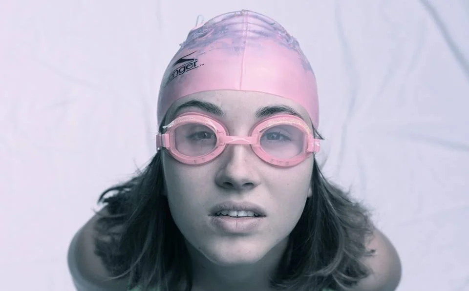 Close-up of a young woman in swimming gear, wearing a pink swim cap and goggles, looking directly at the camera against a neutral background.