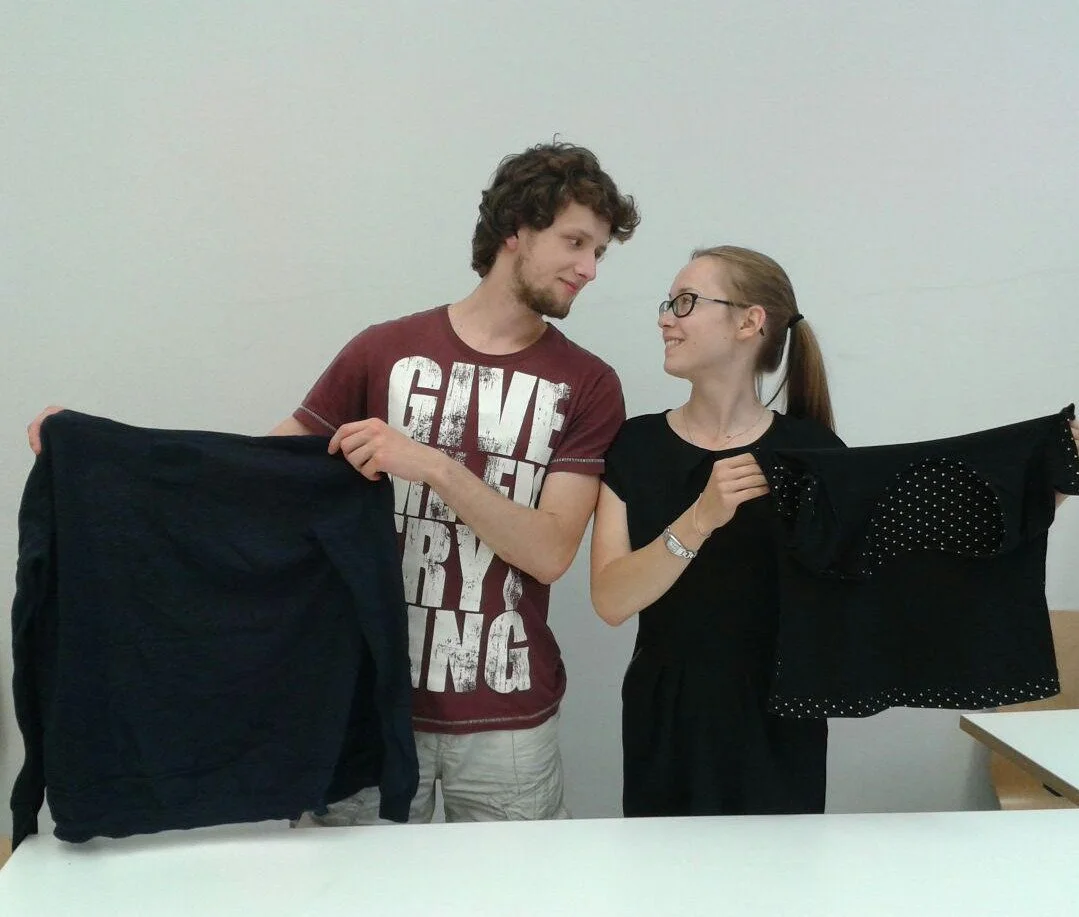 A young man and a young woman standing indoors, holding up black clothing items and smiling at each other.