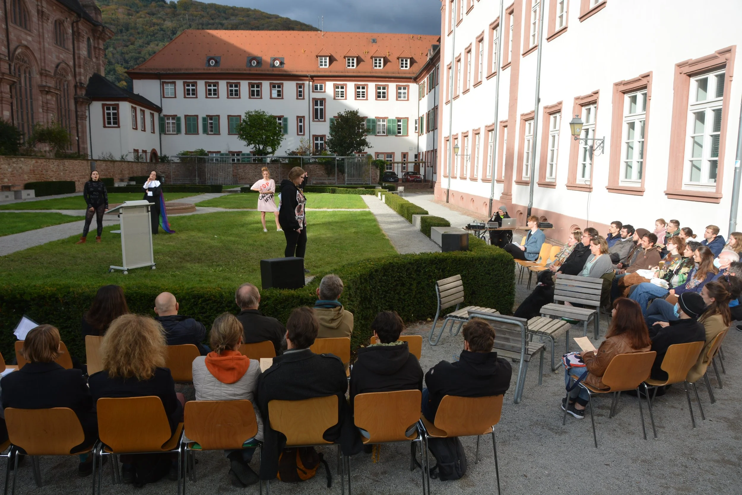Outdoor theatrical performance with actors on a small stage in a garden, audience seated in chairs, some audience members taking notes or photos, and a person operating a keyboard.