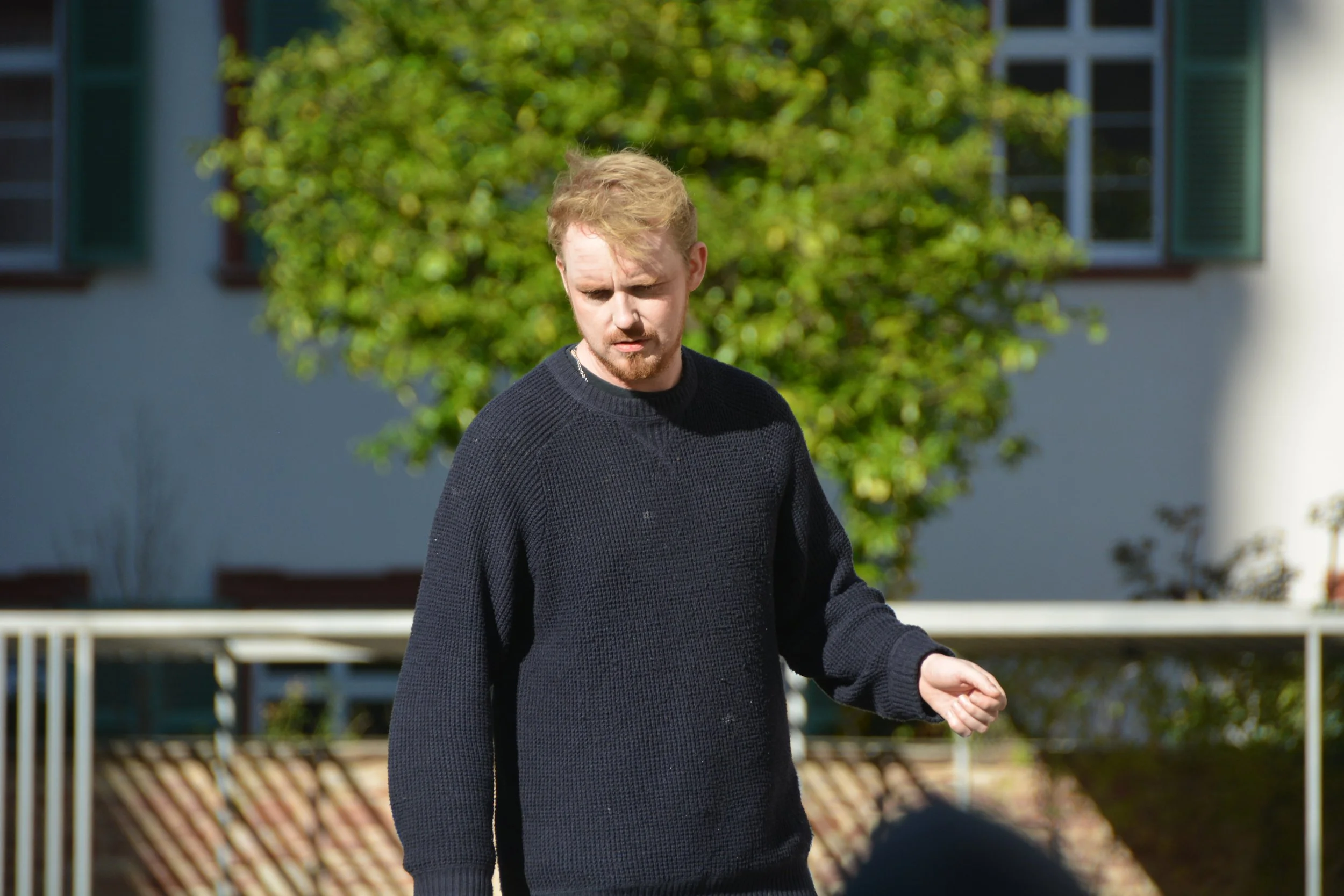 A young man with light-colored hair and a beard wearing a black sweater standing outdoors on a sunny day, with green bushes and residential buildings in the background.