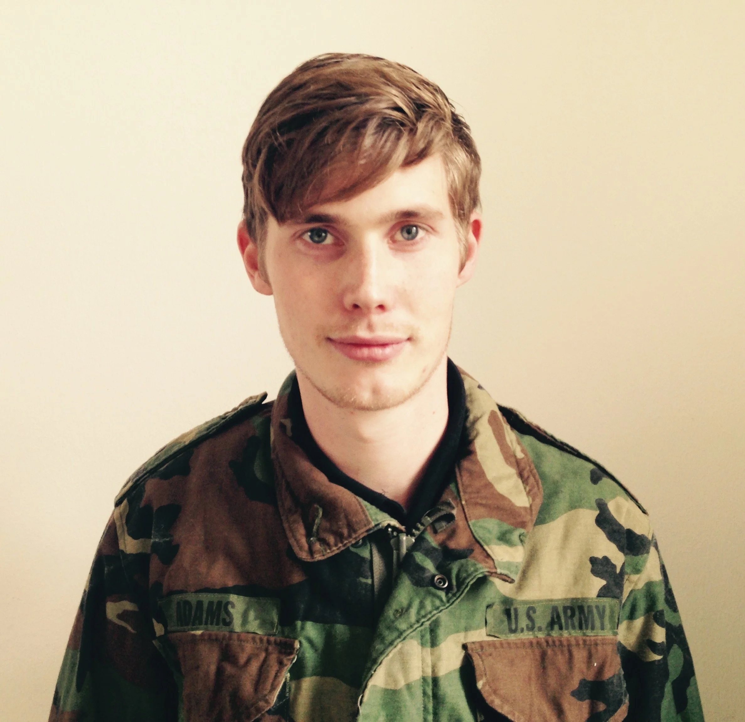 A young man in U.S. Army camouflage uniform standing against a plain light-colored wall.