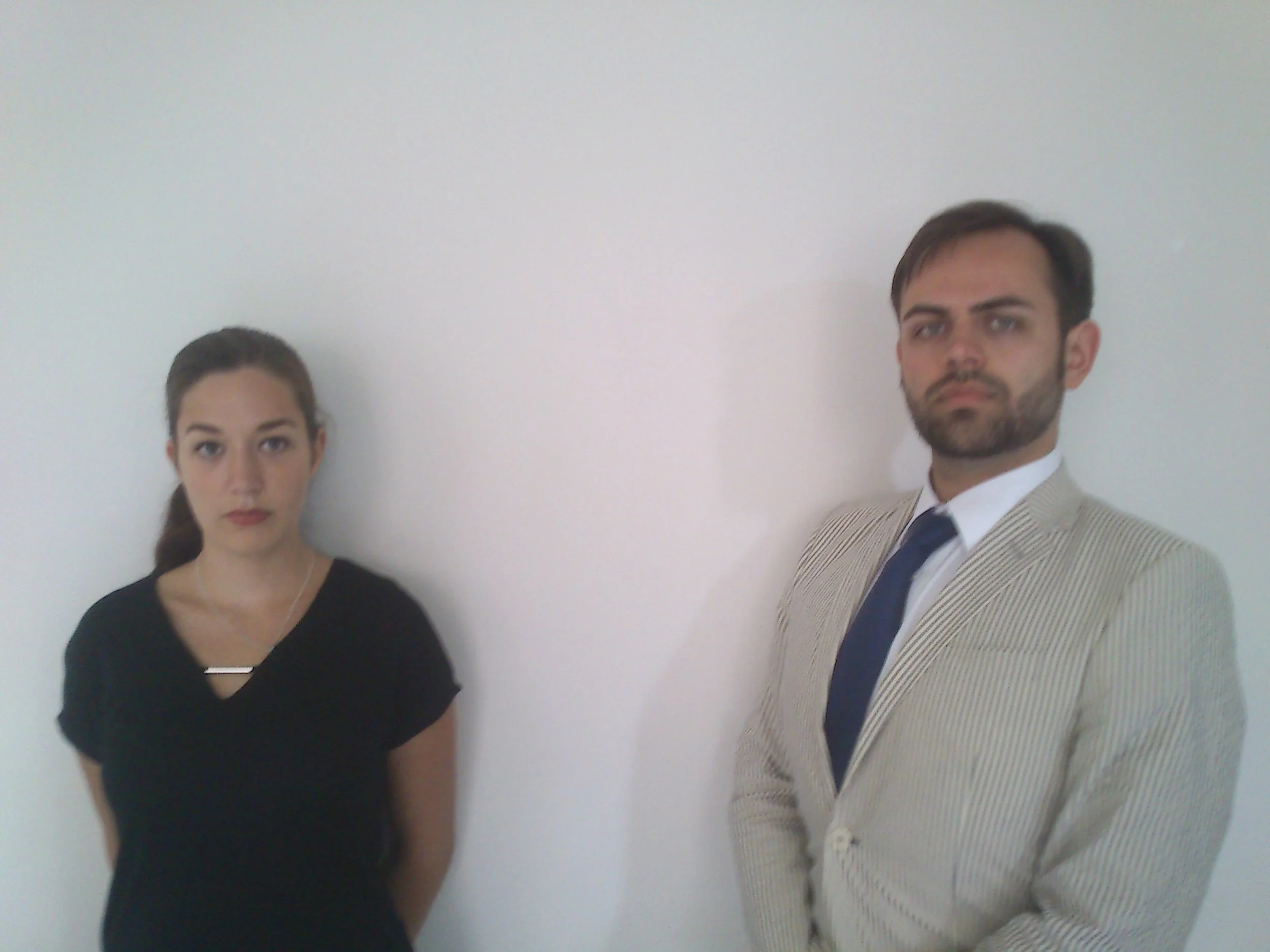 A woman and a man standing against a plain white wall. The woman has long dark hair, and is wearing a black top with a necklace. The man has dark hair, facial hair, and is dressed in a light-colored striped suit with a white shirt and dark tie.
