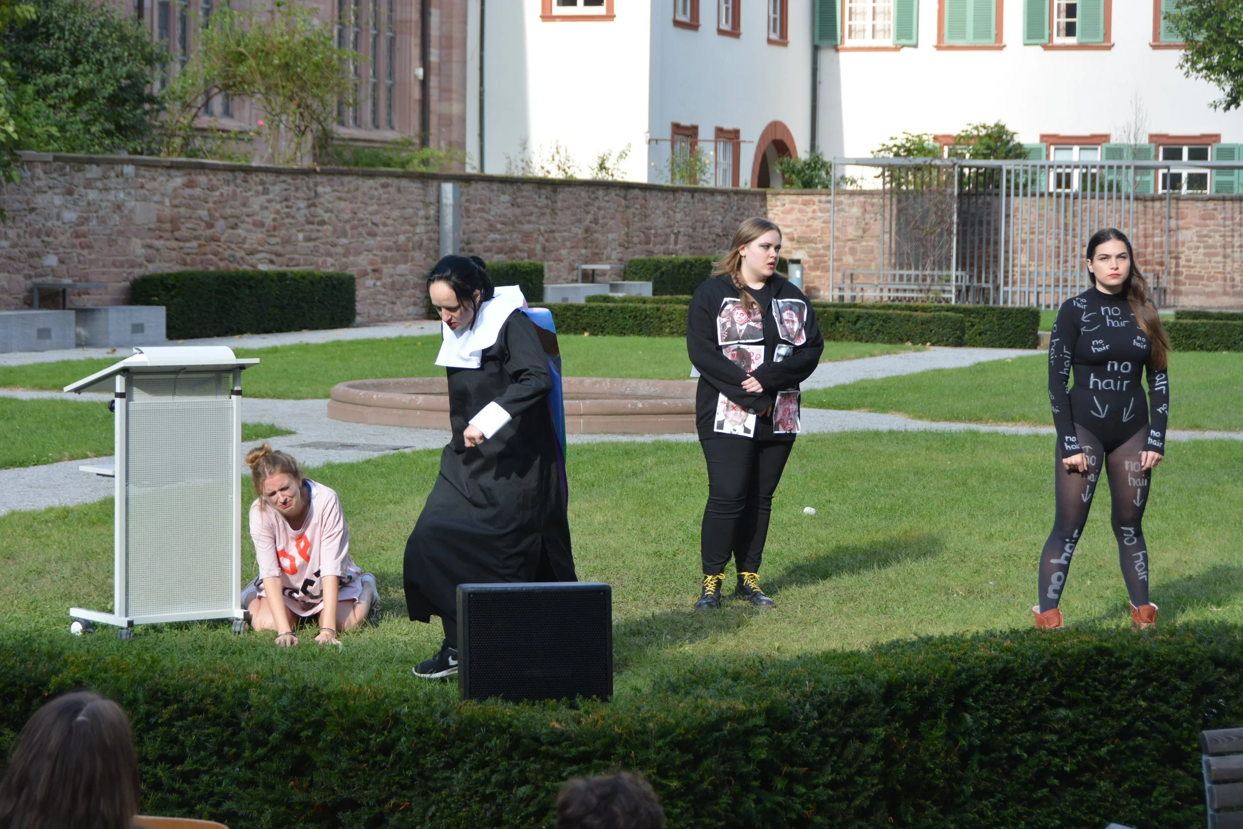 Four women are on a grassy outdoor stage. One is kneeling on the ground, looking distressed. The second woman, dressed in a black gown, is standing and looking down. The third woman, wearing a black hoodie with images on it, stands with her arms crossed. The fourth woman, in a black bodysuit with "no hair" written on it all over, stands with her arms at her sides. The audience is partly visible at the bottom of the image.