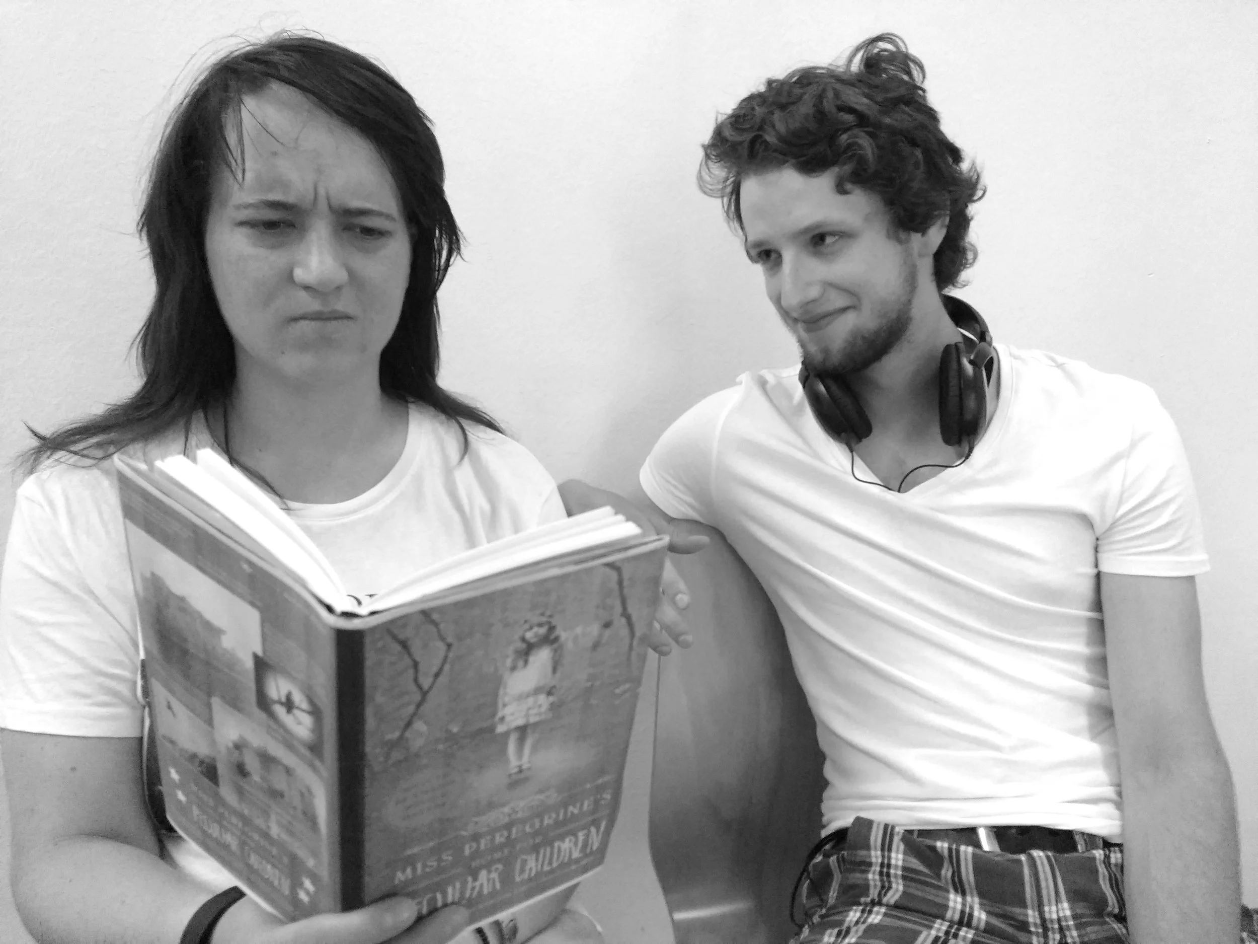 A young woman with a serious facial expression reading a book, seated next to a young man with curly hair and headphones, smiling slightly, against a plain wall.