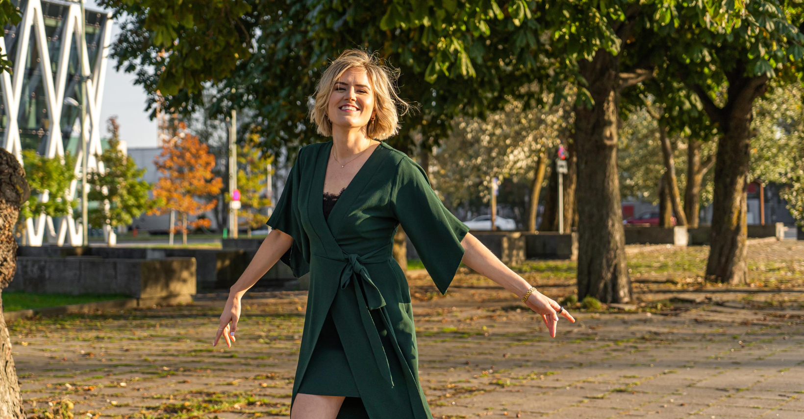 A woman with blonde hair wearing a dark green dress with a black undershirt, standing outdoors in a park with trees and autumn foliage, smiling and posing with her arms extended.