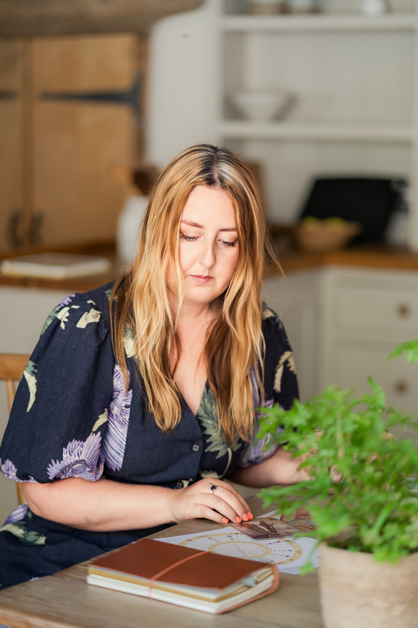 A woman with long blonde hair, wearing a dark floral shirt, sits at a wooden table reading a magazine, with a notebook and a potted plant nearby in a cozy kitchen setting.