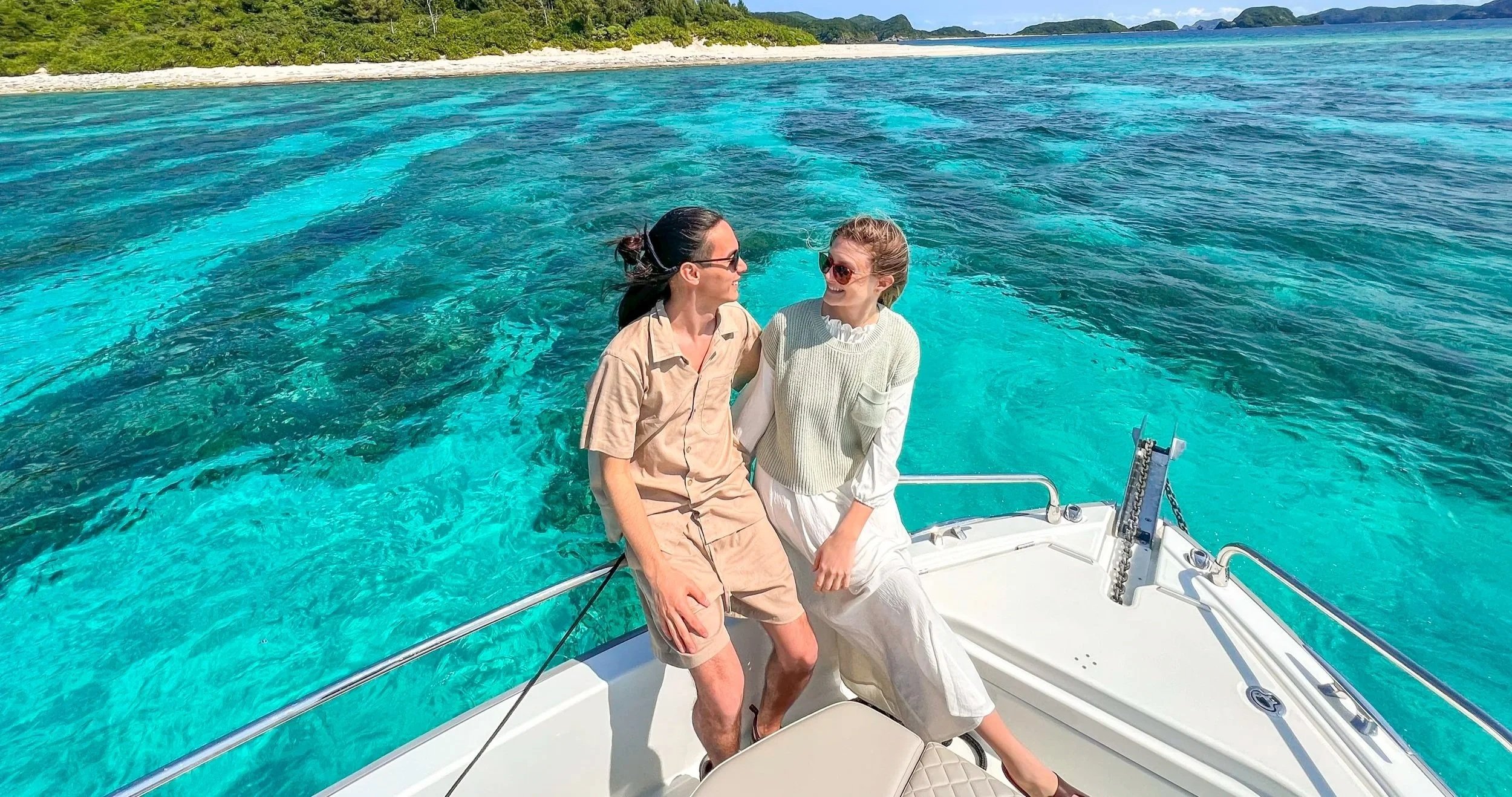 青い海と白い砂浜のあるリゾート地で、二人の女性が白いボートに座って笑いながら会話している