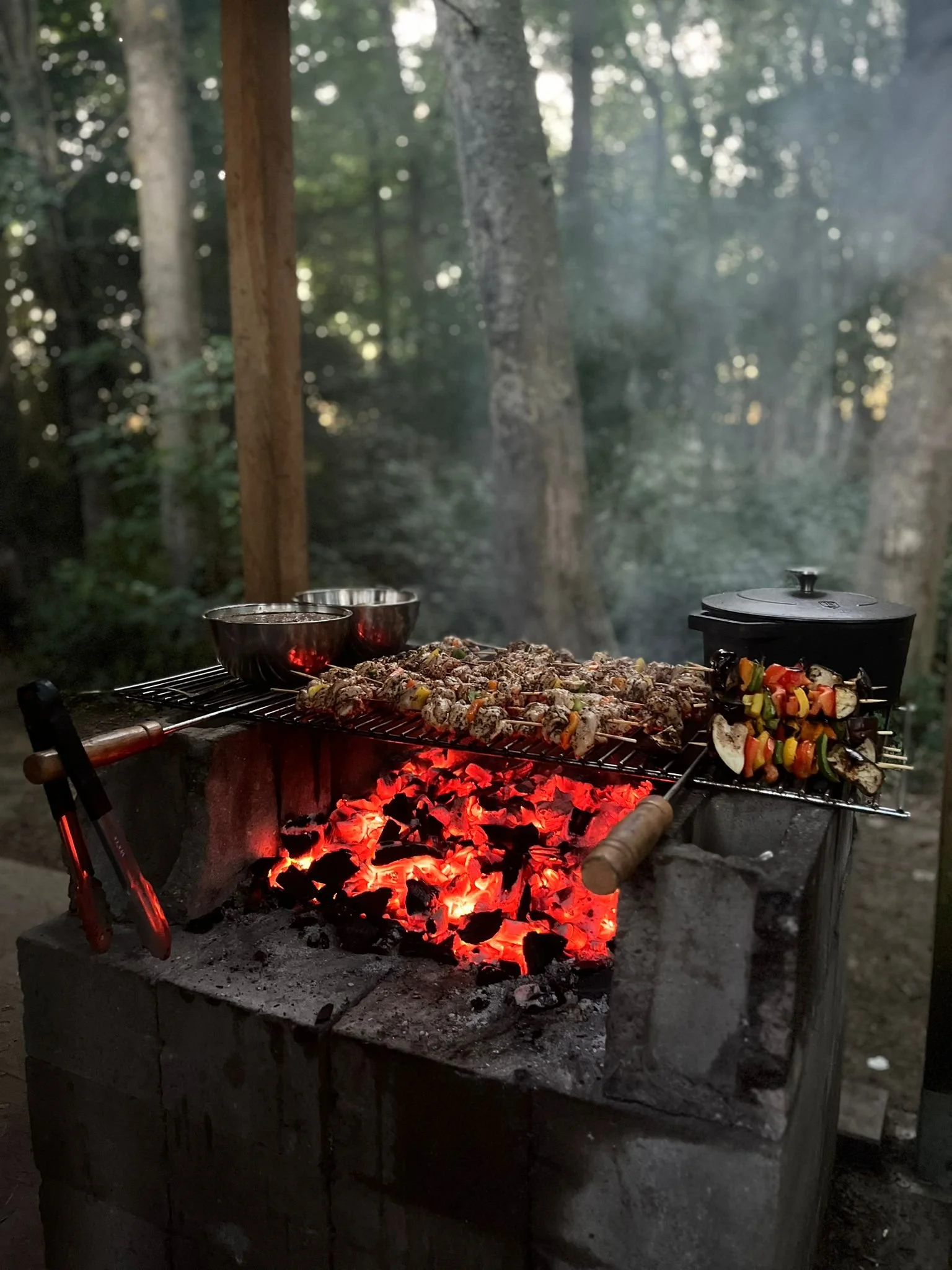 Barbecue en plein air dans une forêt avec nourriture grillée, notamment des brochettes et des plats en sauce, sur une grille au-dessus de braises rouges.