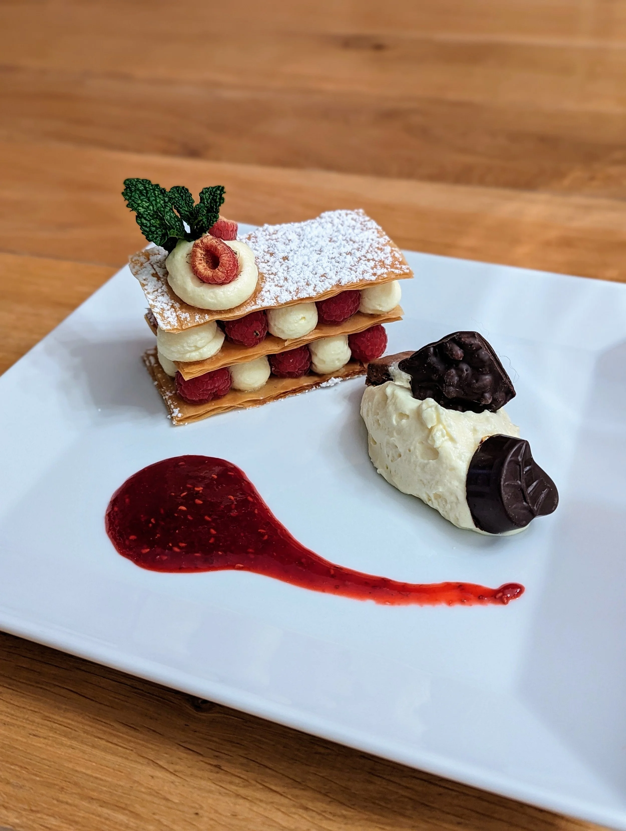 Dessert avec mille-feuille à la crème, fruits rouges et feuille de menthe, accompagné de sorbet à la vanille avec morceaux de chocolat, et sauce aux fruits rouges sur une assiette blanche.