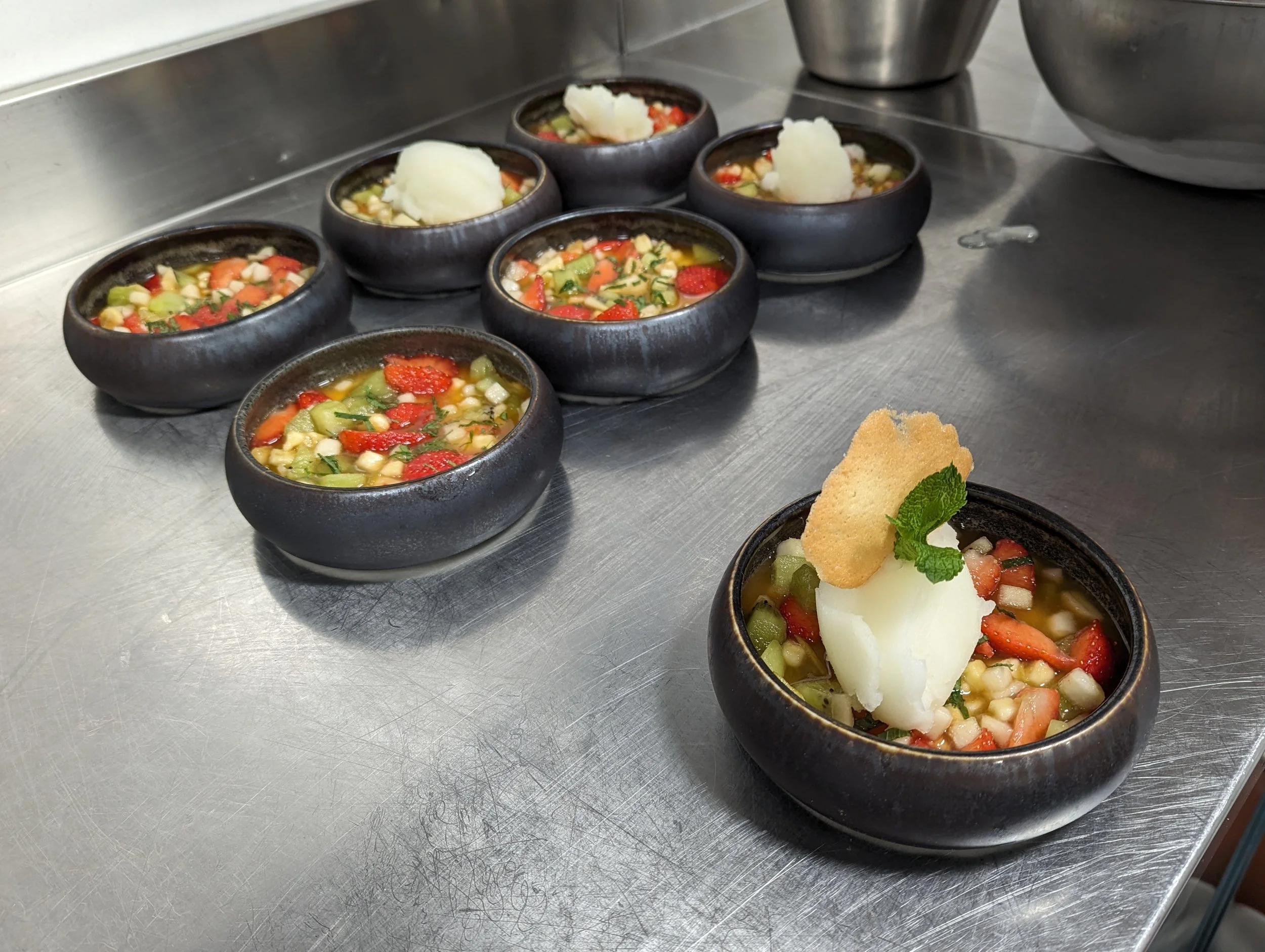 Petites portions de soupe de légumes dans des bols en céramique noire, garnies de purée de pommes de terre, d'une feuille de menthe et d'une tuile croustillante, sur un plan de travail en métal.