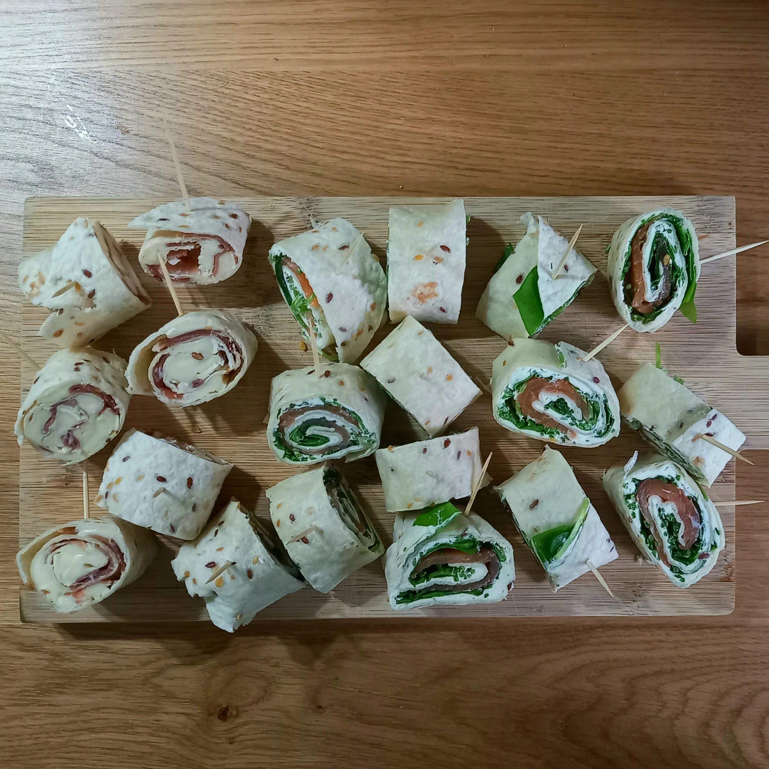 Rouleaux de wrap avec divers ingrédients, présentés sur une planche en bois. Formes variées, contenant des légumes et du jambon, piqués avec des cure-dents.