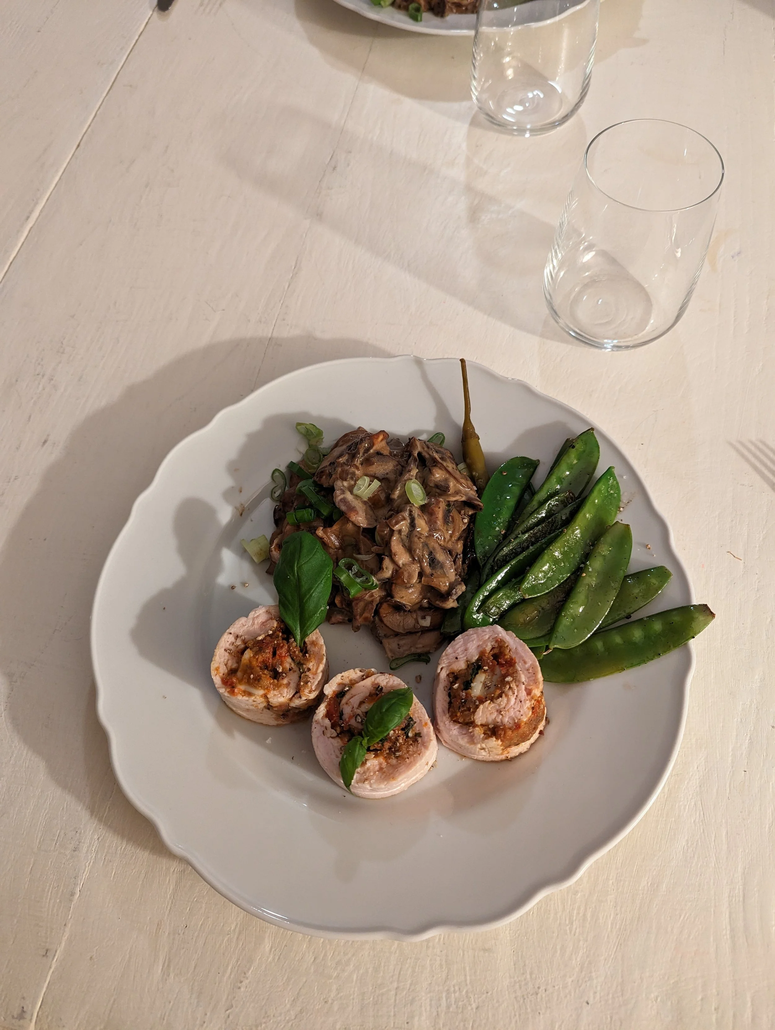Assiette de viande enroulée de bacon avec sauce, haricots verts, salade de champignons, et garniture de basilic, sur une table en bois clair, assortiment de verres vides à proximité.