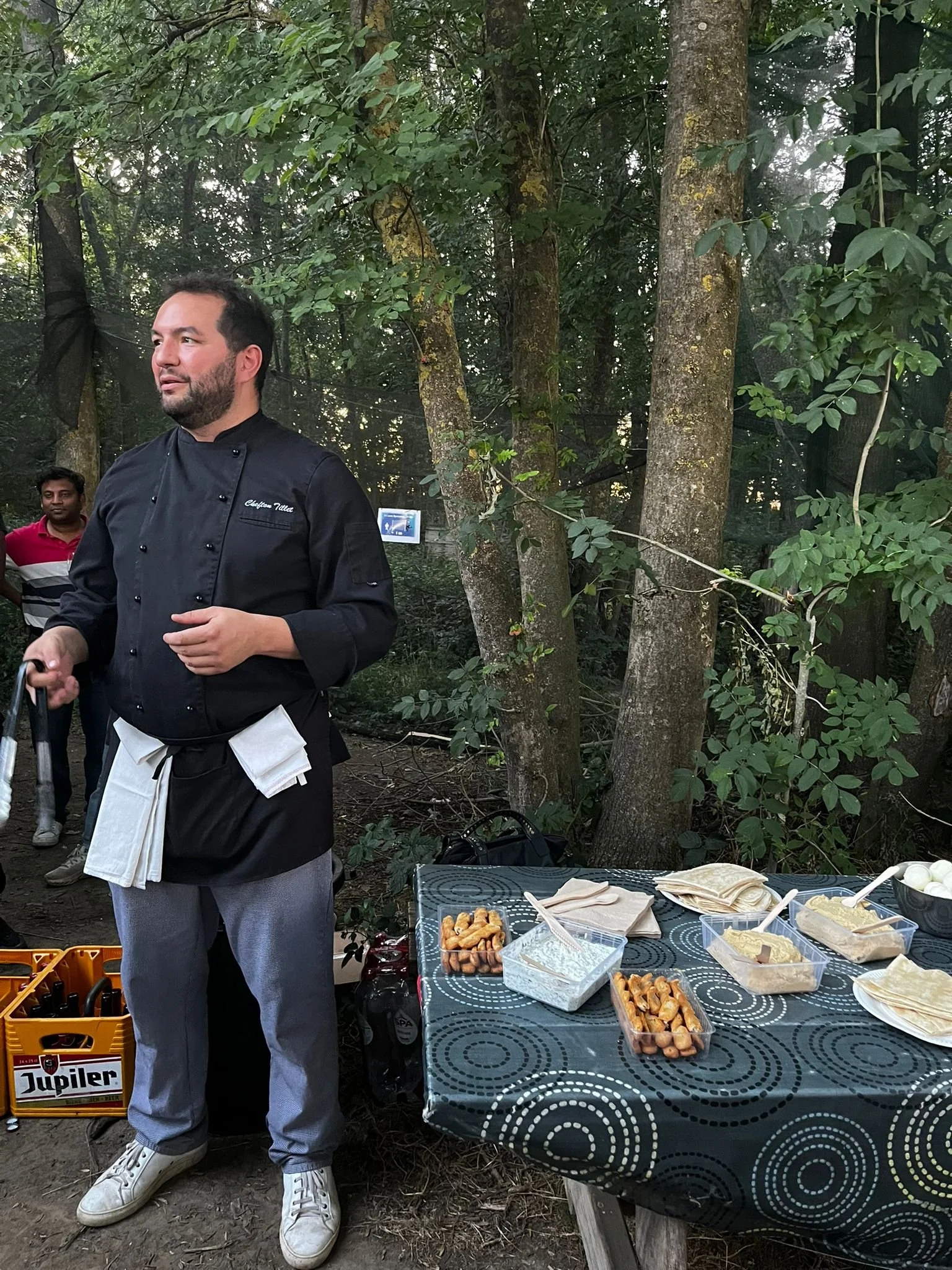 Chef en uniforme dans un environnement forestier avec une table contenant des aliments comme des sauces, des saucisses, des tortillas et des œufs