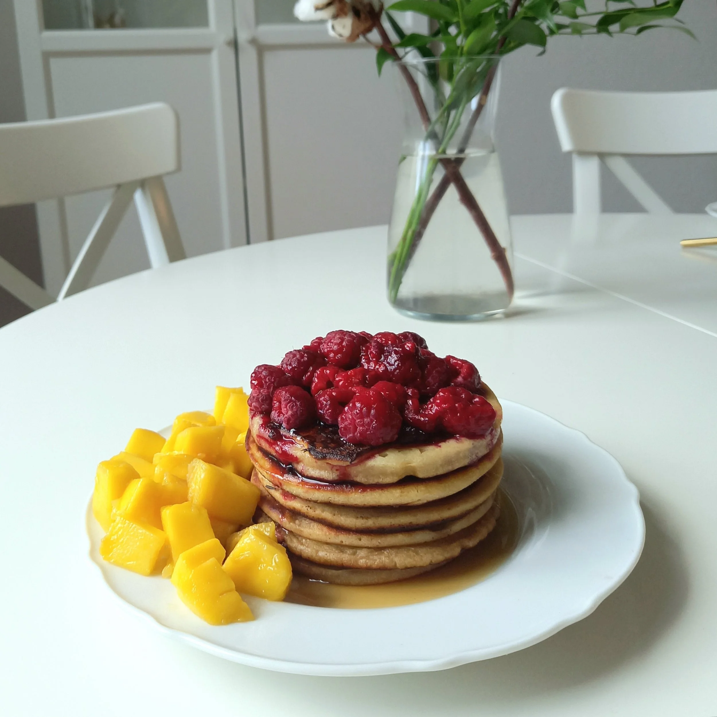 Une assiette de pancakes garnis de framboises et de sirop, accompagnée de morceaux de mangue, sur une table blanche avec une vase contenant des fleurs en arrière-plan.