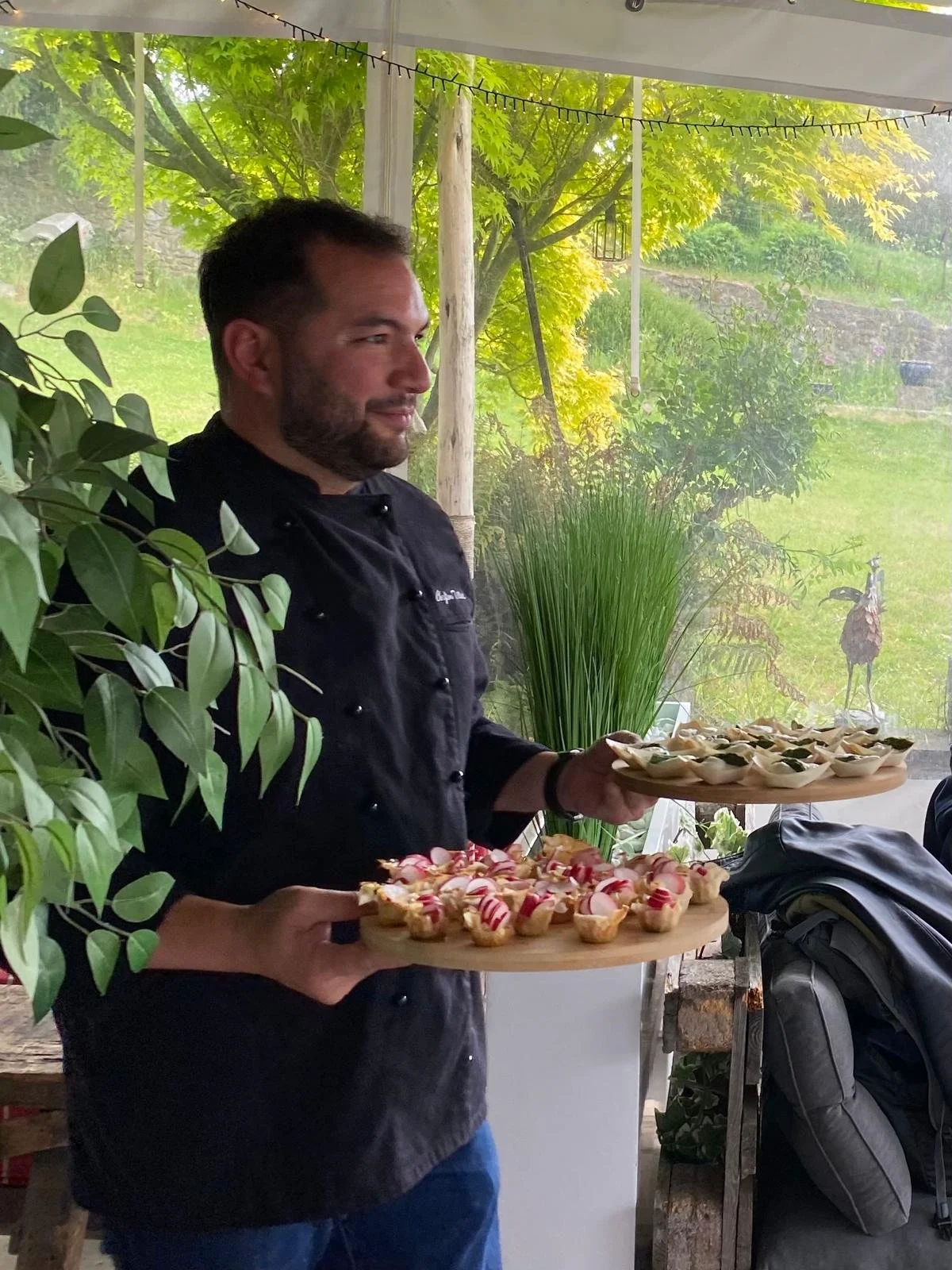 Un chef souriant tient deux plateaux avec des amuse-bouches, dans un espace avec beaucoup de végétation et de lumière naturelle.
