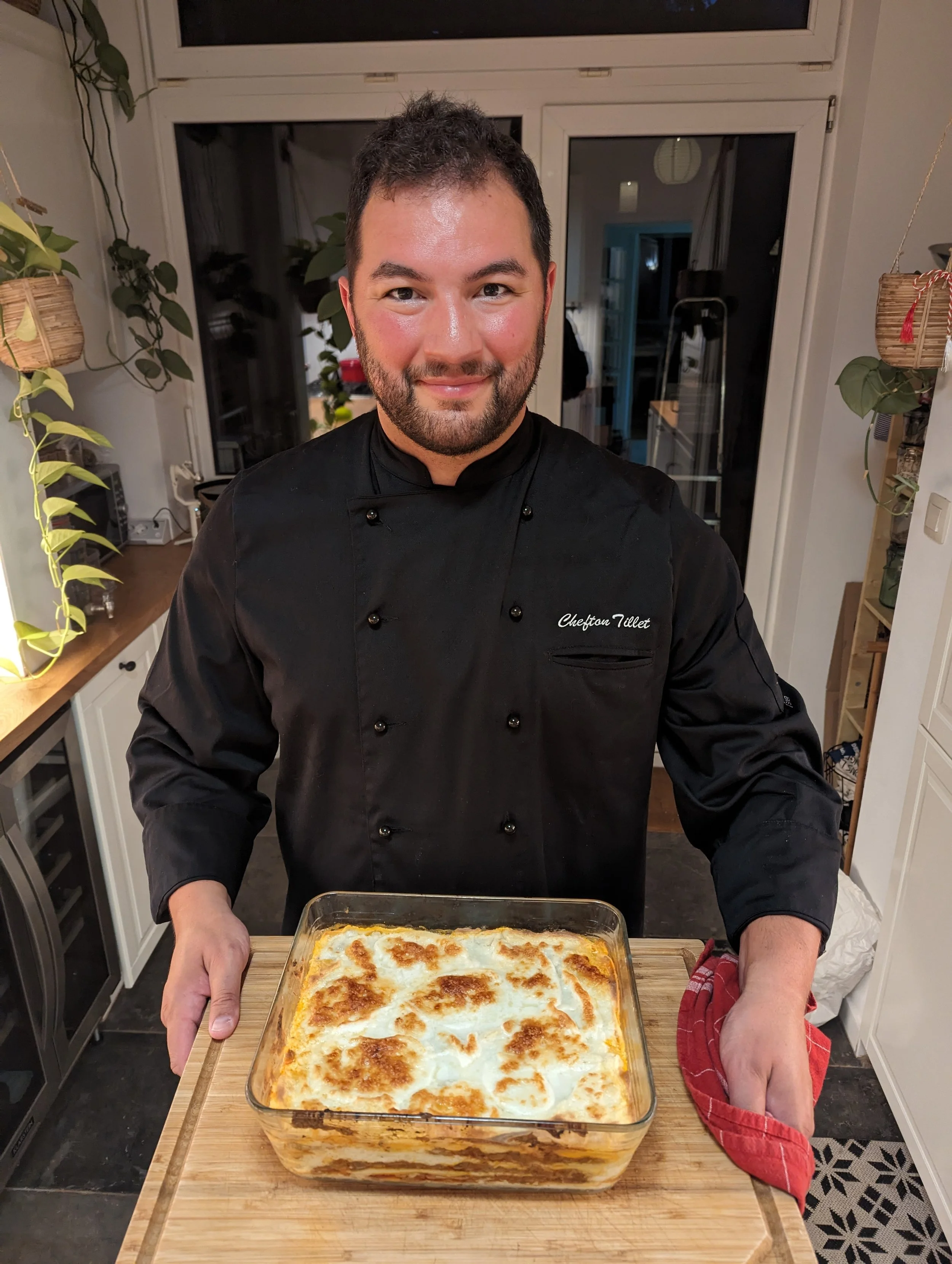 Homme en costume de chef cuisinier présentant un plat de lasagnes cuites en gratin dans un plat en verre, dans une cuisine moderne.