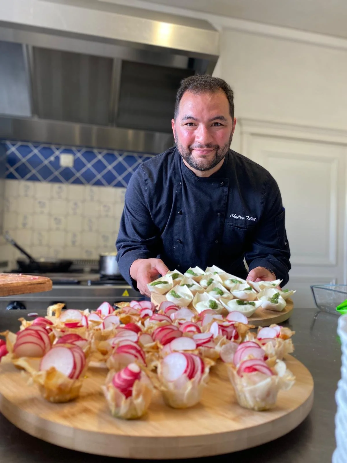 Un chef en tenue professionnelle présente une assiette de petits amuse-bouches ou tapas sur une table, avec d'autres petits amuse-bouches en forme de cuplets garnis de radis et autres garnitures.