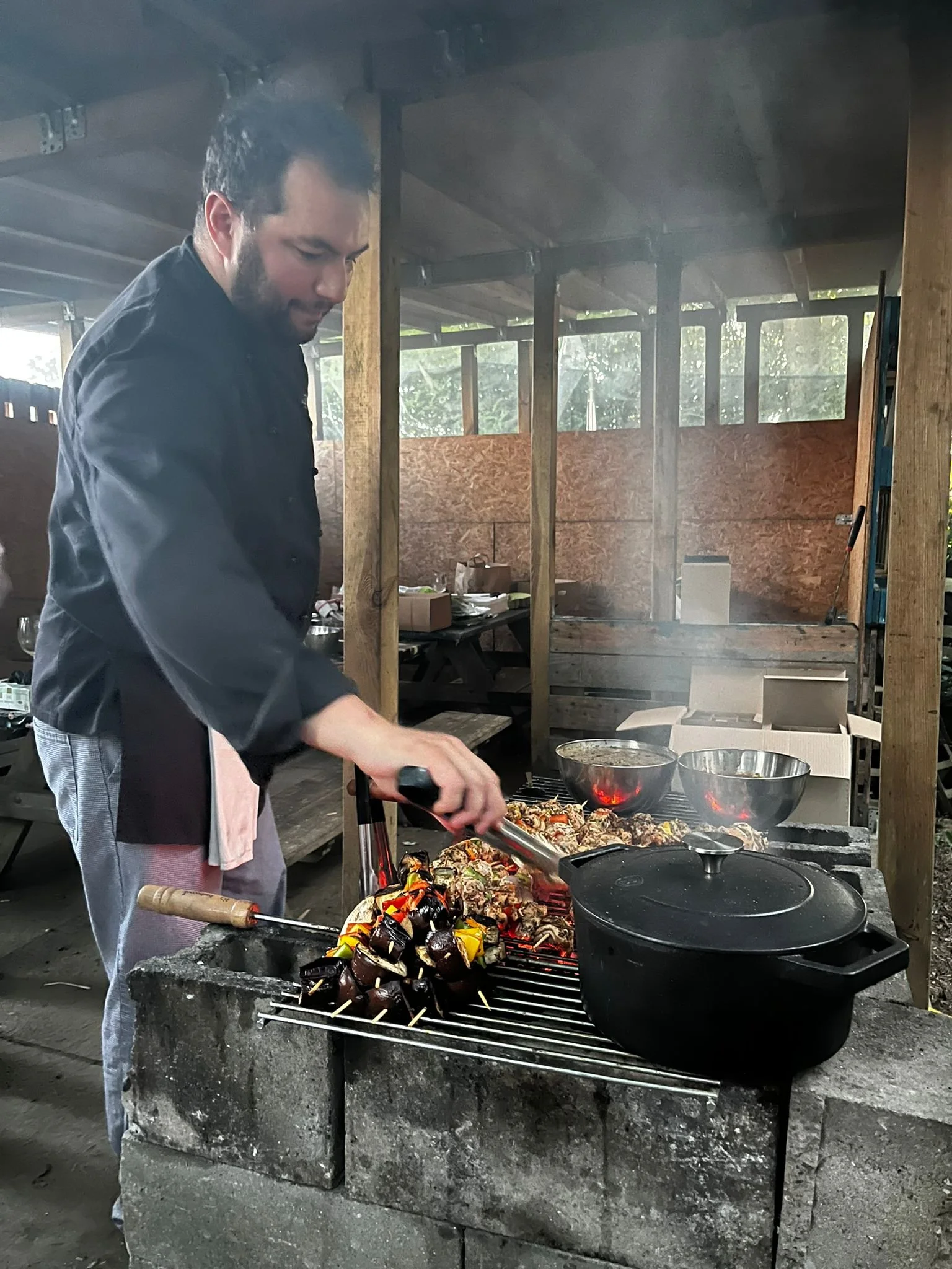 Un homme cuisine des brochettes de légumes et de viande sur un barbecue extérieur en bois.