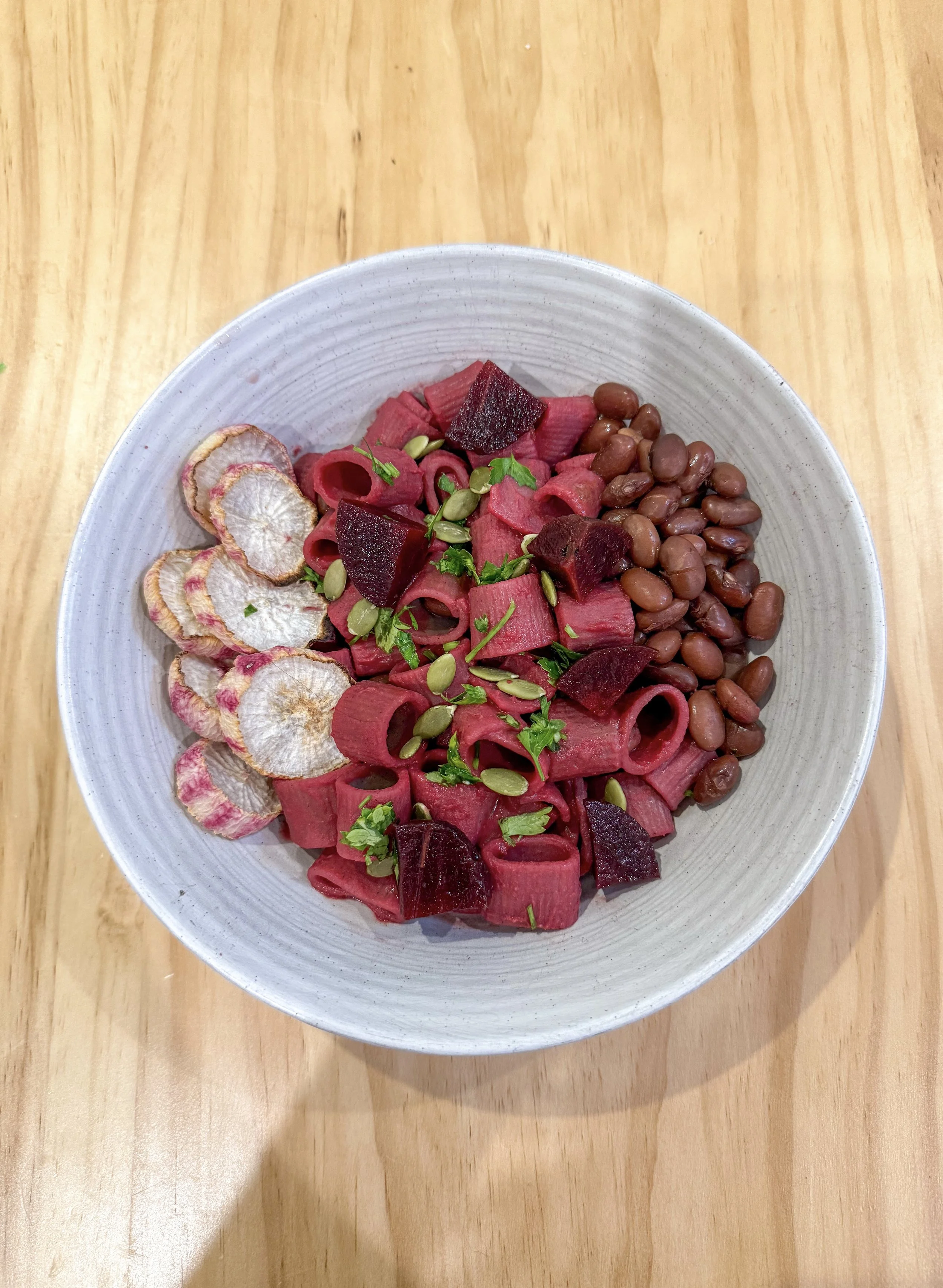 Bowl of beetroot pasta salad with beans, dried radish chips, and herbs on a wooden table.