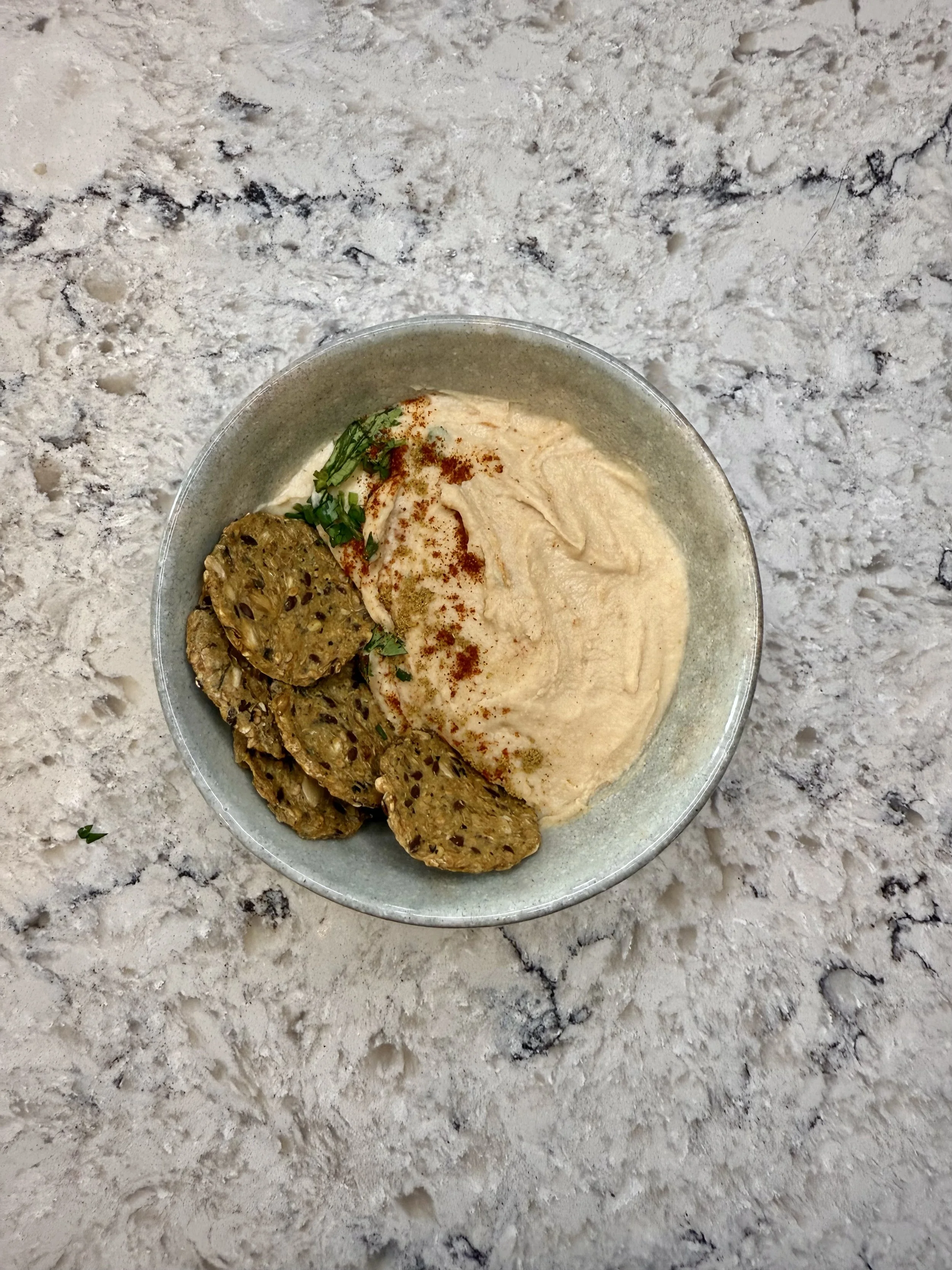 A bowl of hummus garnished with paprika, parsley, and chickpea slices, placed on a marble countertop.