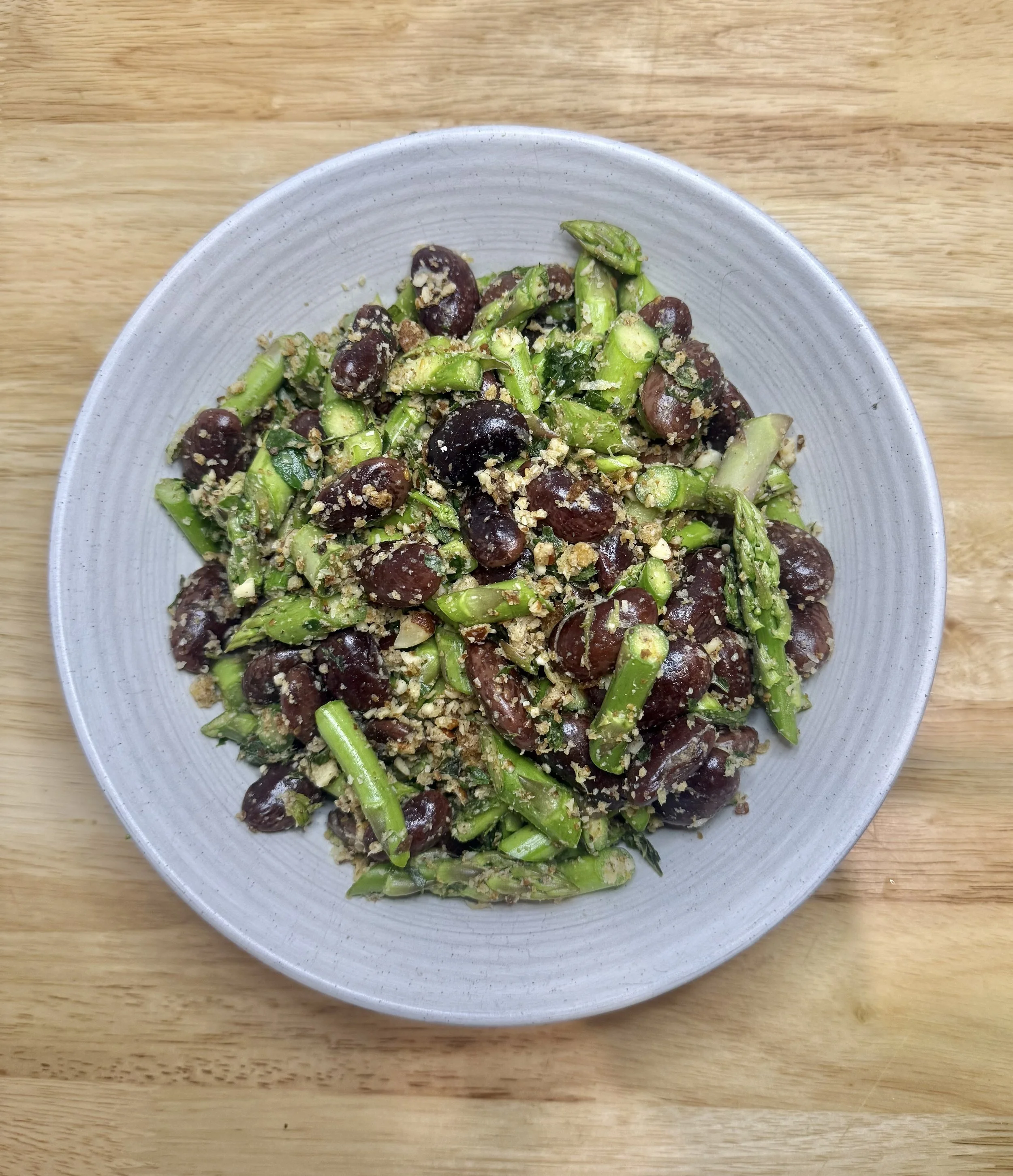 A bowl of cooked kidney beans and asparagus spears with crushed nuts or breadcrumbs on top, on a wooden table.