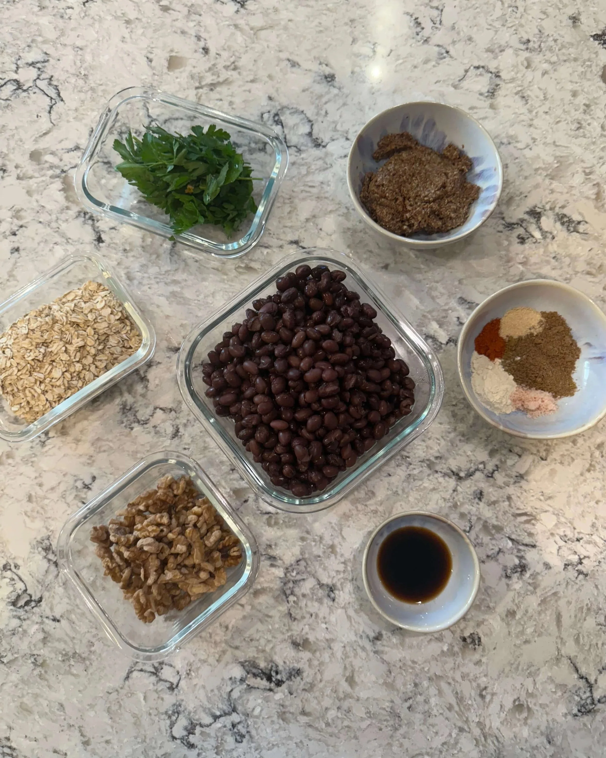 Ingredients for black bean burgers including black beans, oats, walnuts, ground flaxseed, parsley, spices, and tamari on a kitchen counter.