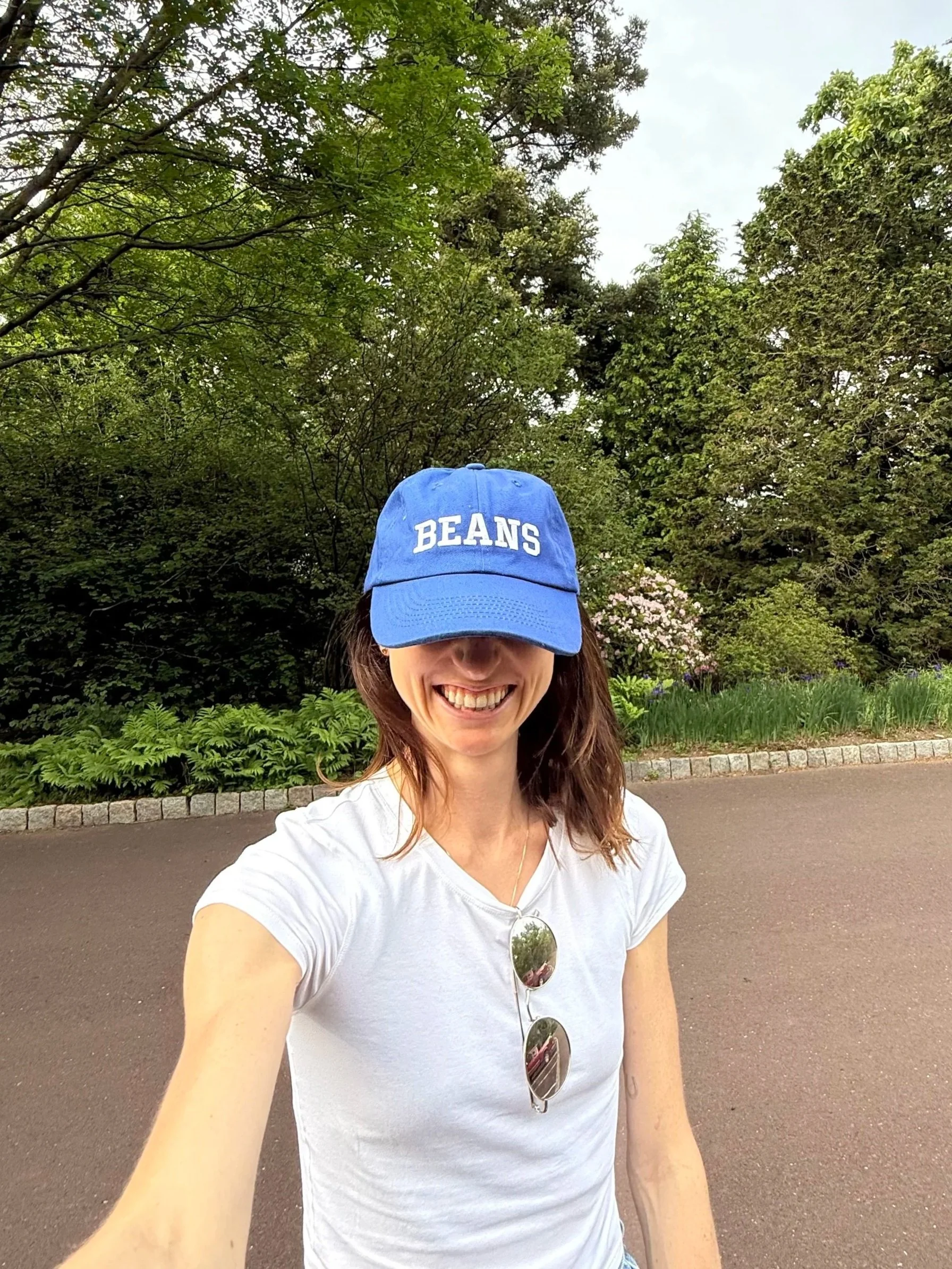 A smiling woman wearing a white t-shirt, sunglasses hanging from her neck, and a blue cap with the word "BEANS" on it, taking a selfie outdoors on a paved pathway with trees and bushes in the background.