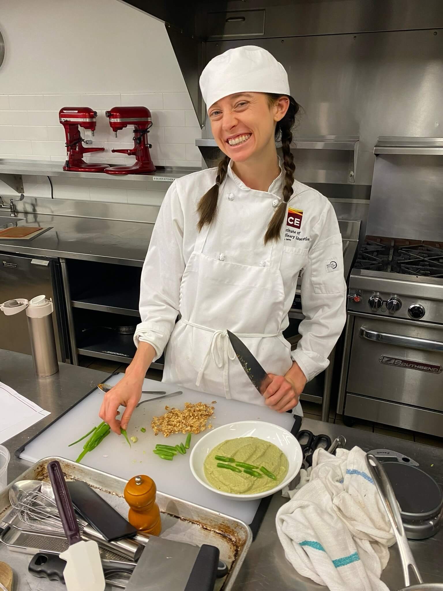 A woman in a chef uniform and hat is smiling and standing in a commercial kitchen, preparing food on a cutting board with chopped nuts, green beans, and mashed avocado or guacamole in a white bowl.
