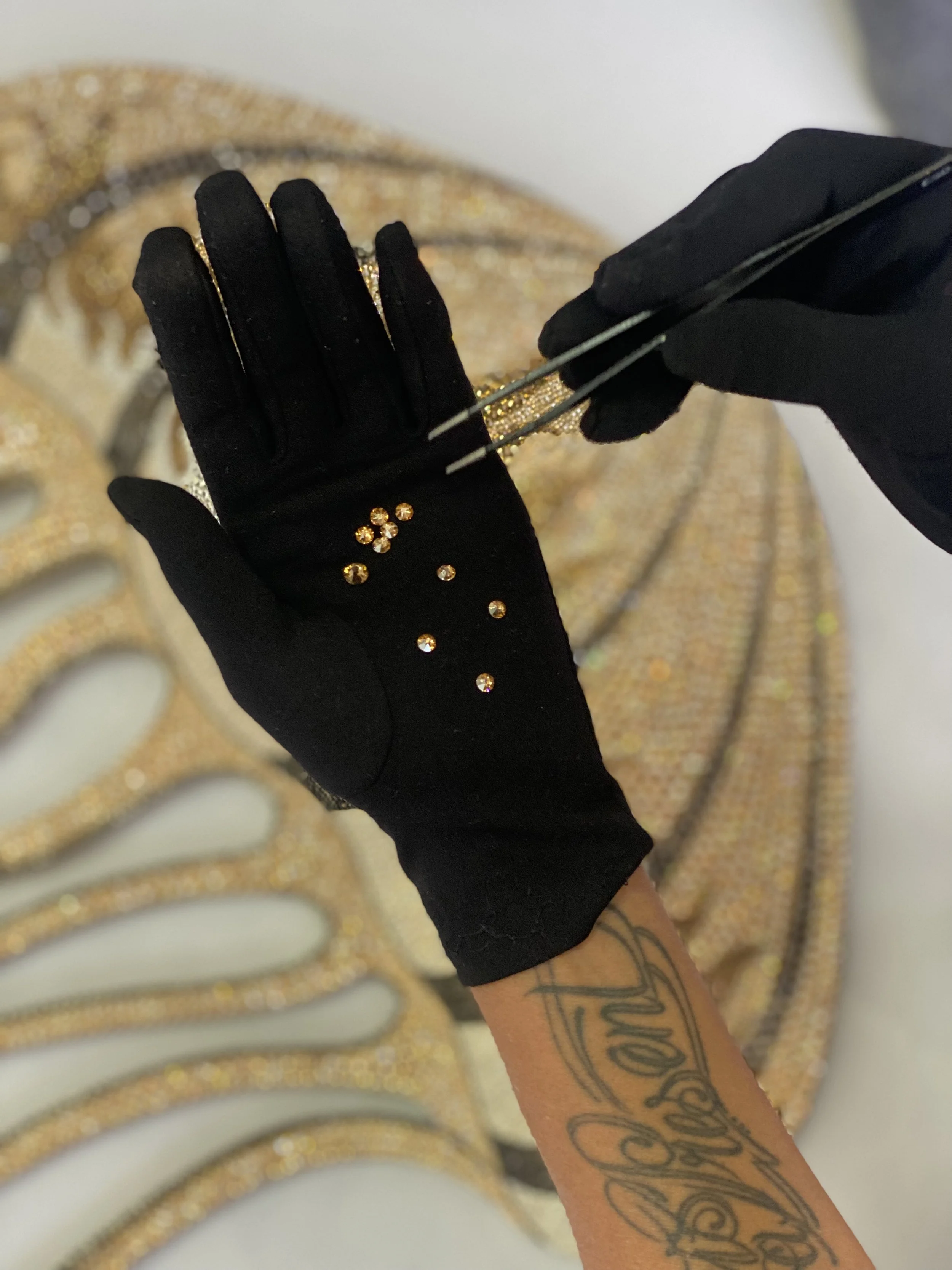 A person wearing black gloves is placing small yellow gemstones onto a golden, ornate jewelry piece with tweezers. The jewelry is decorated with many more yellow gemstones arranged in patterns.
