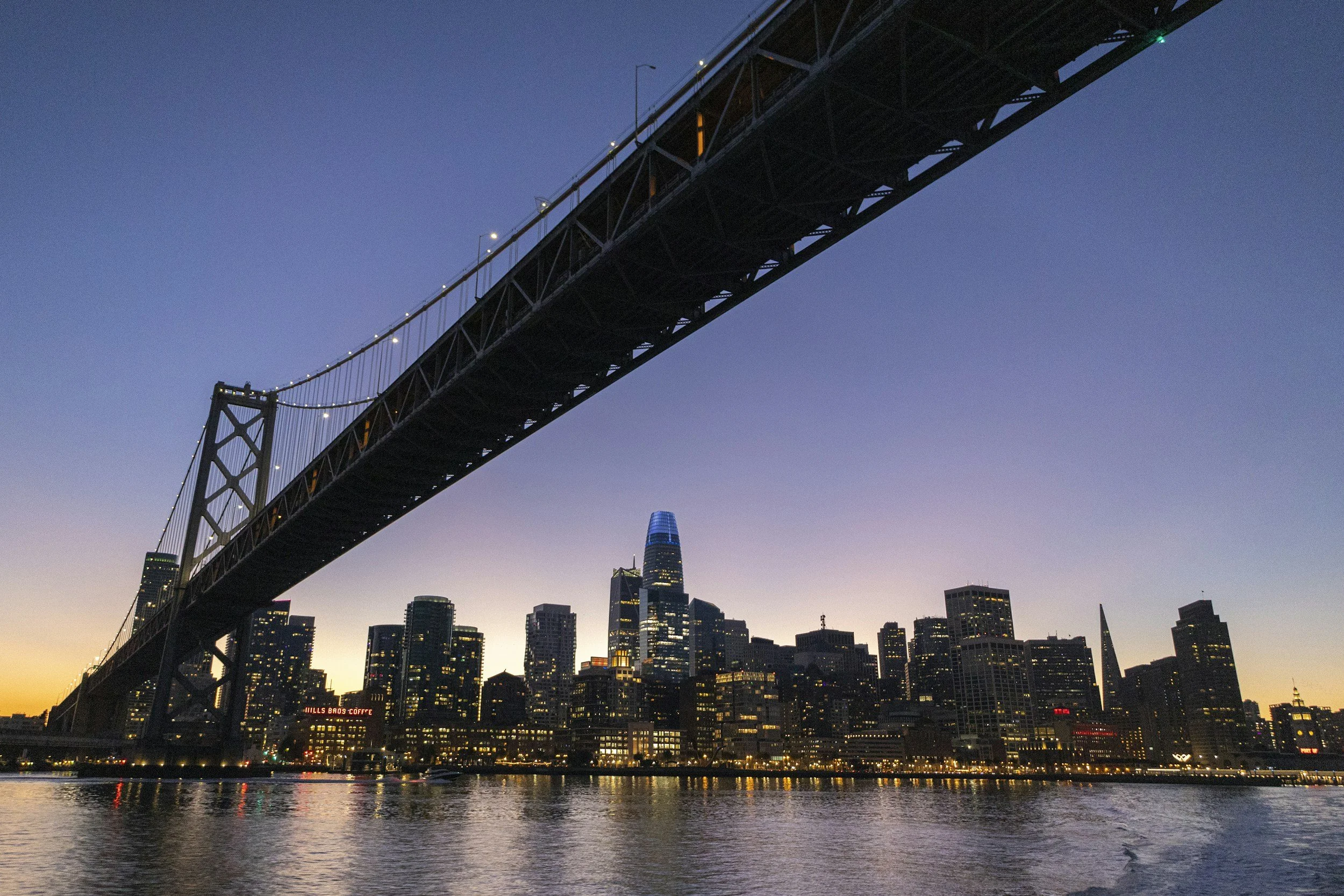 City skyline at dusk with a large bridge in the foreground and tall buildings with illuminated windows in the background. The water below reflects the city lights.