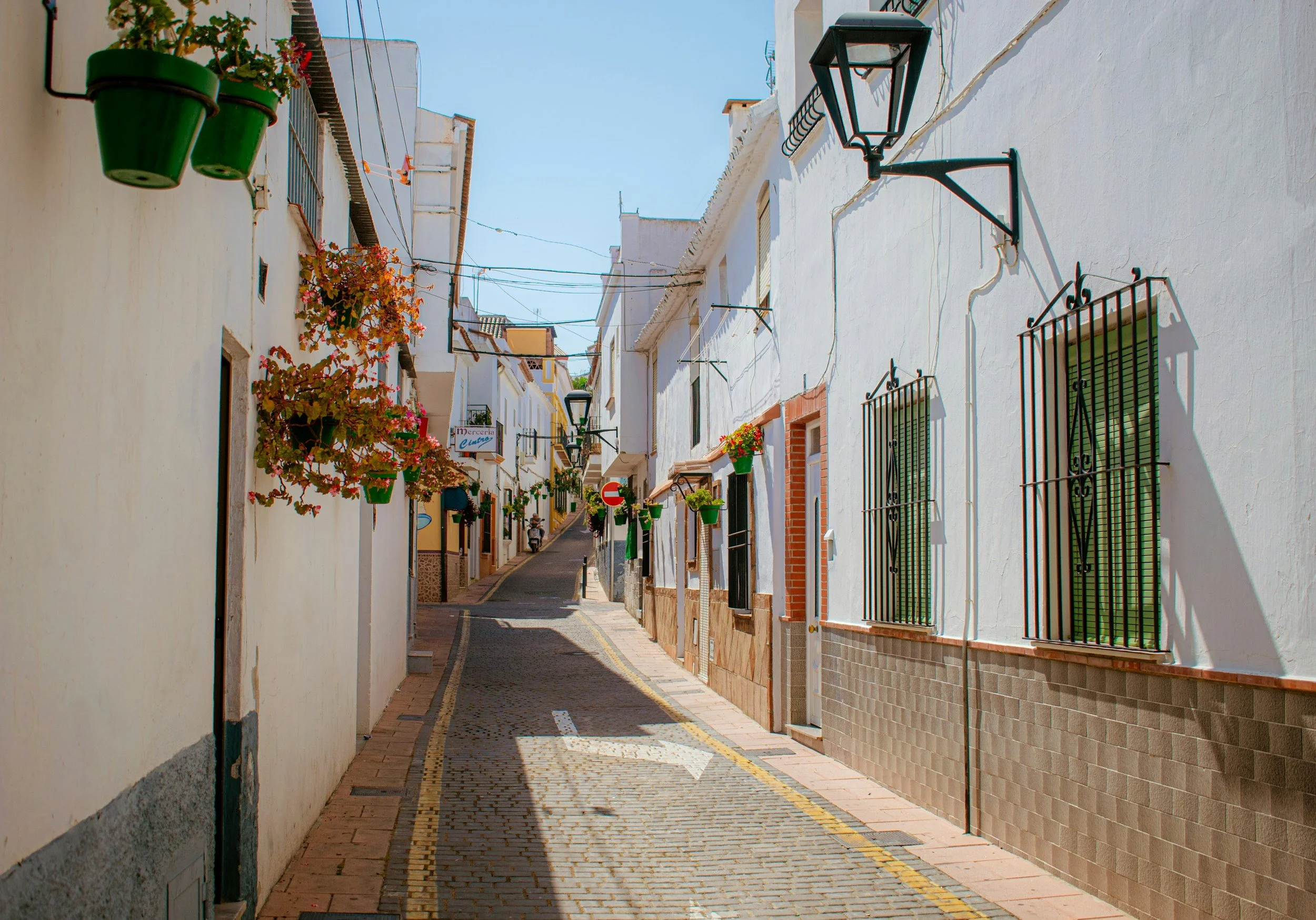 A narrow Spanish street with white buildings, balconies with flowers, black iron window grilles, and street lamps under a clear blue sky.