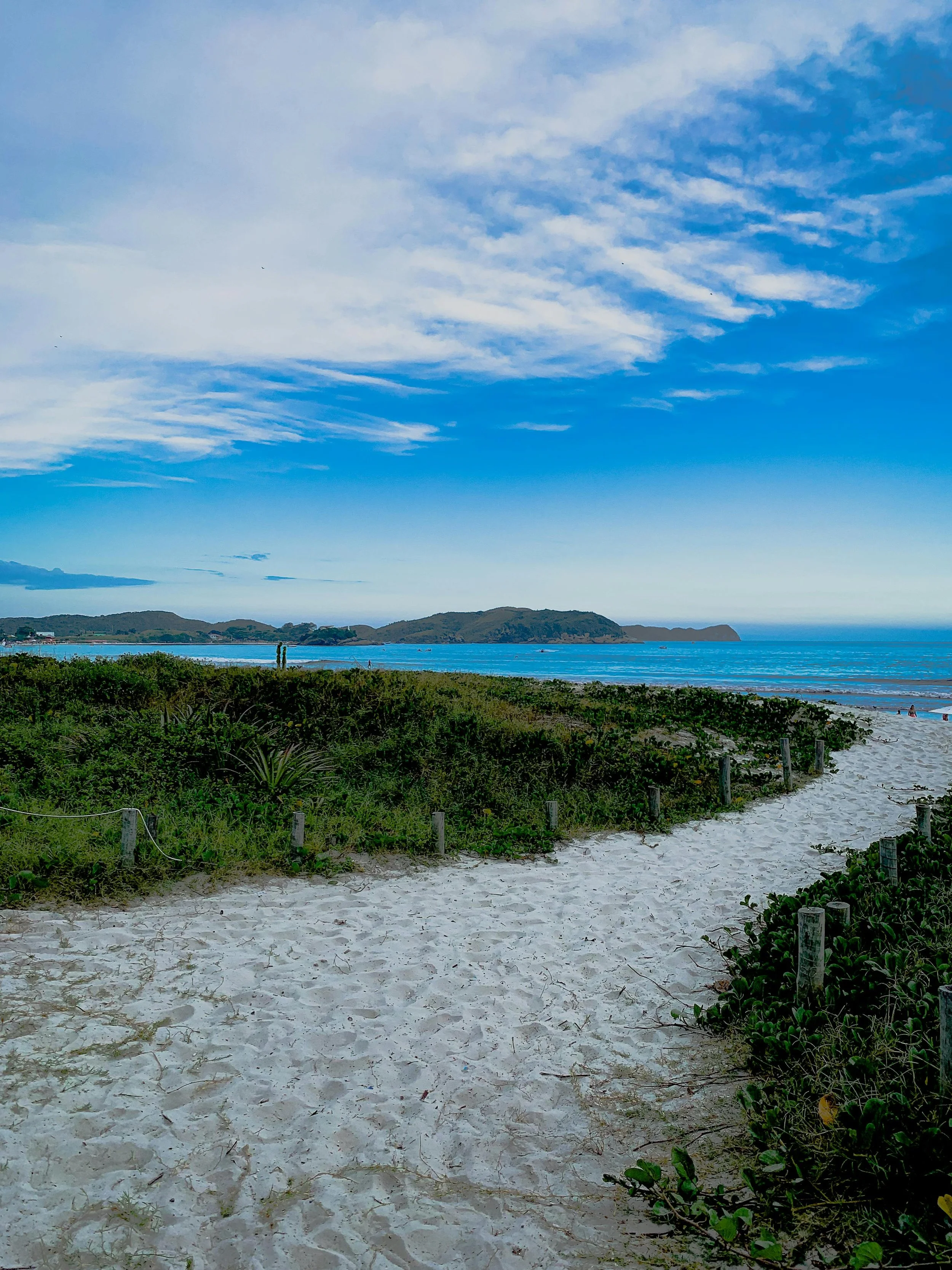 Sandy beach path in Brazil leading to the ocean with green shrubs on either side and a hilly island in the distance under a partly cloudy blue sky.