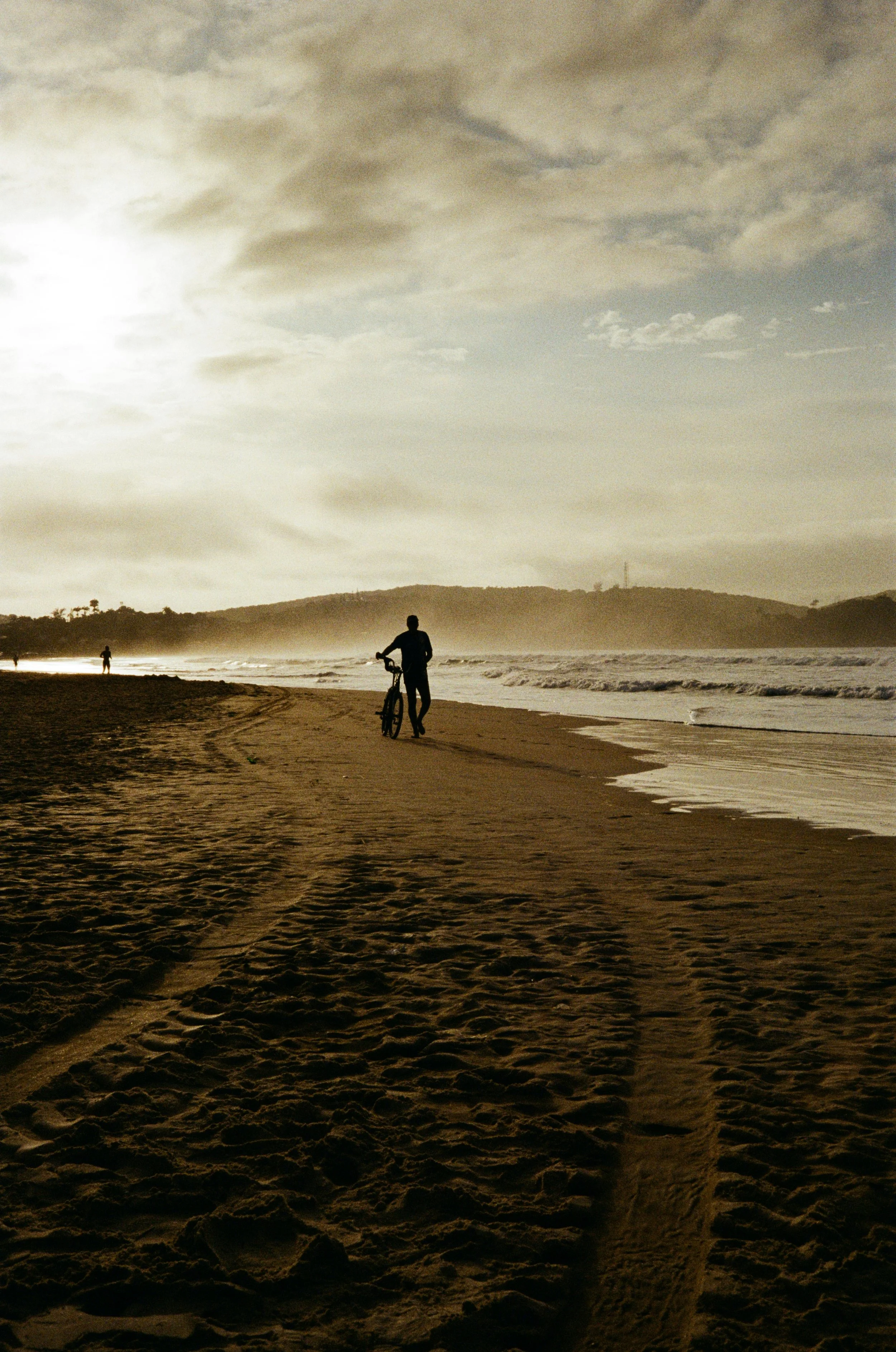 Person walking a bicycle on a beach in Brazil during sunset with a few other people in the distance.