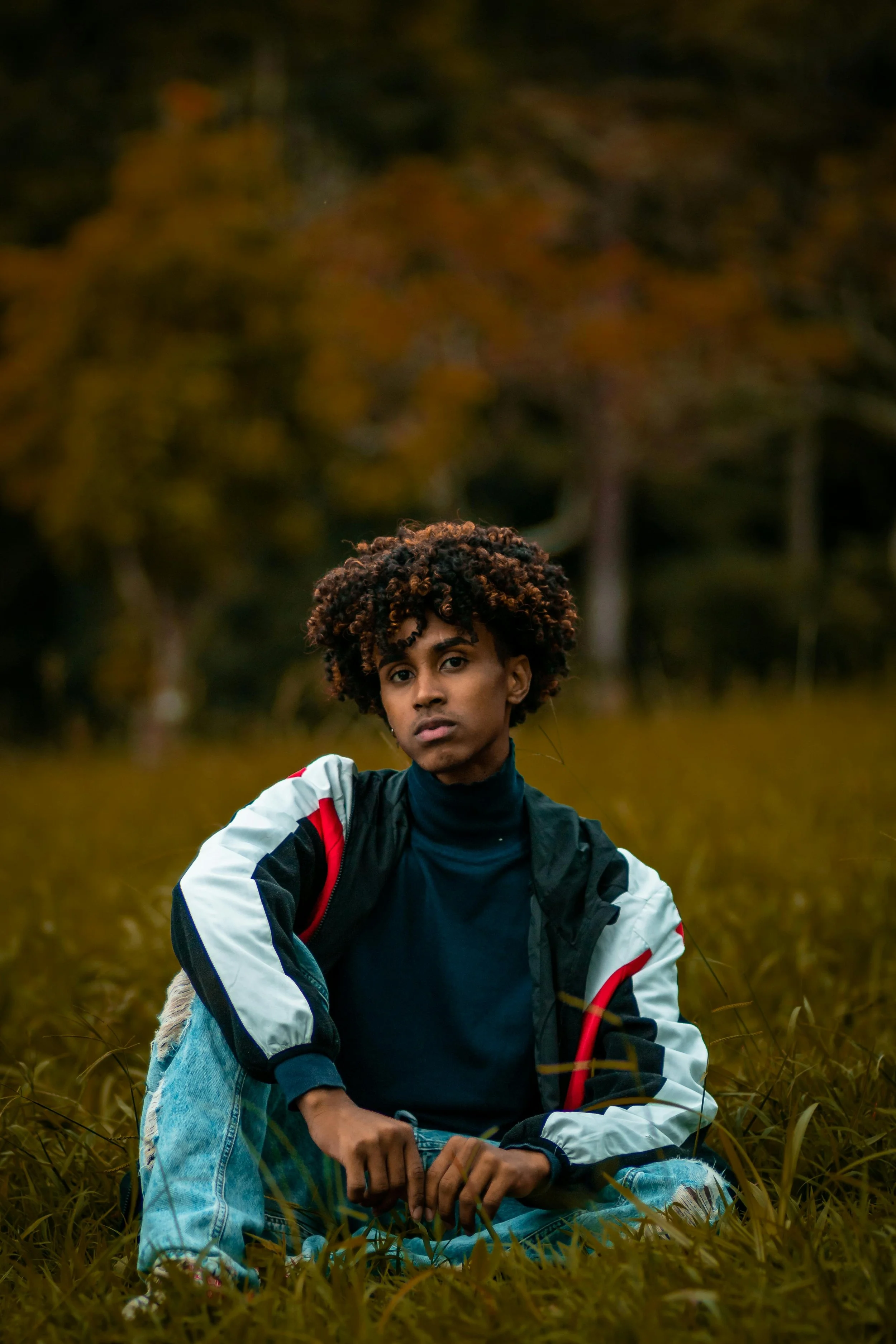 Young Brazilian man with curly hair sitting in a grassy field with autumn trees in the background.