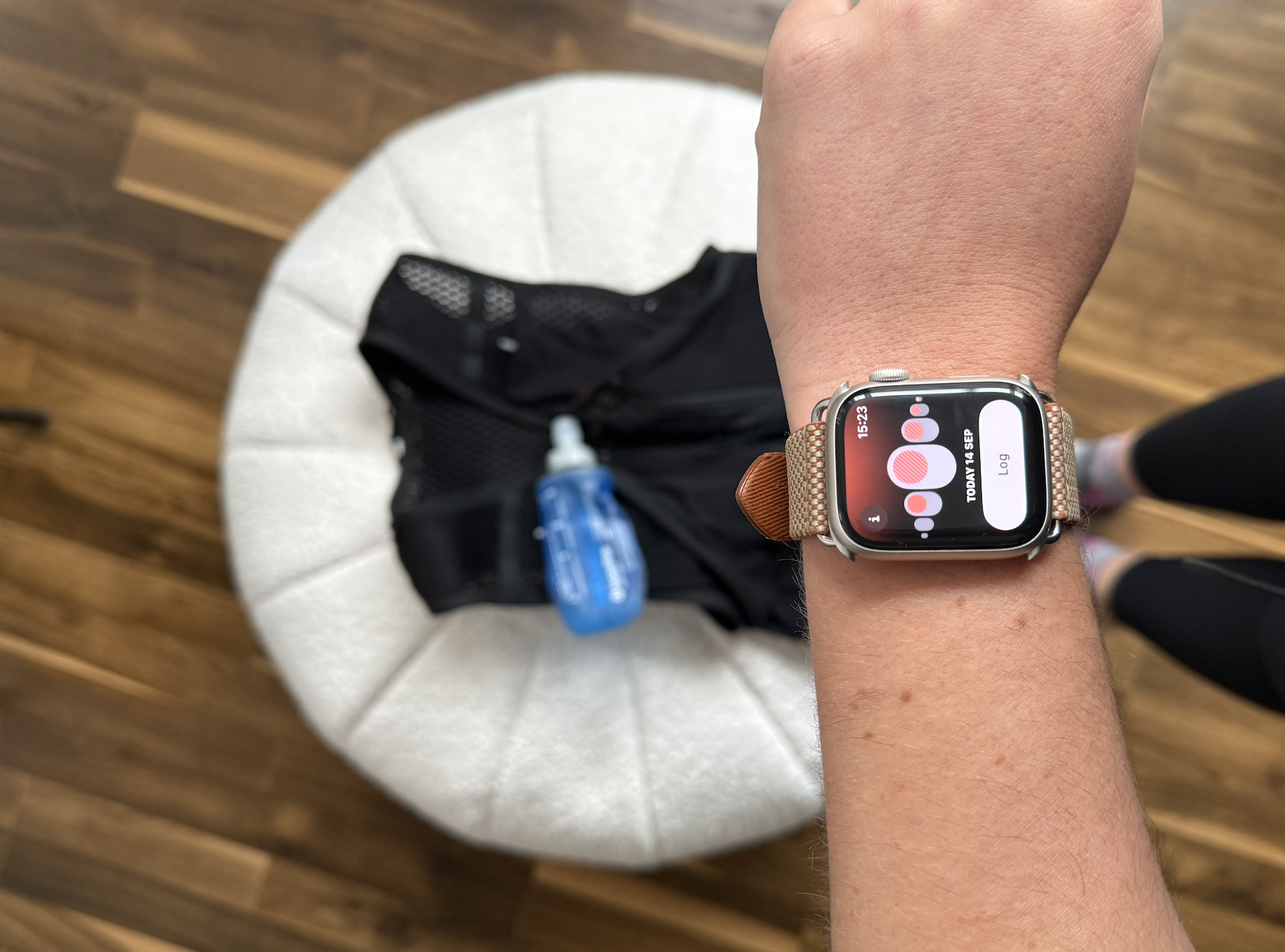 A person's left wrist with an Apple Watch showing a fitness app, positioned above a white circular stool. On the stool, there is a black mesh hydration pack with a blue water bottle attached. The background features a wooden floor and the person's legs in black pants and running shoes.