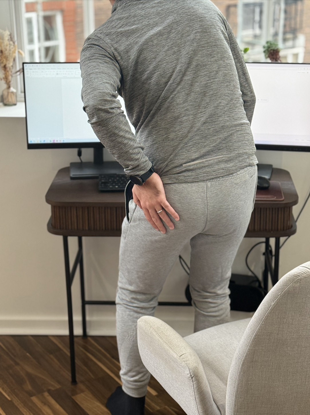 Person wearing a gray sweatshirt and gray sweatpants, standing in front of dual computer monitors on a wooden desk, with one hand on their lower back and the other resting on their hip, in a home office setting.